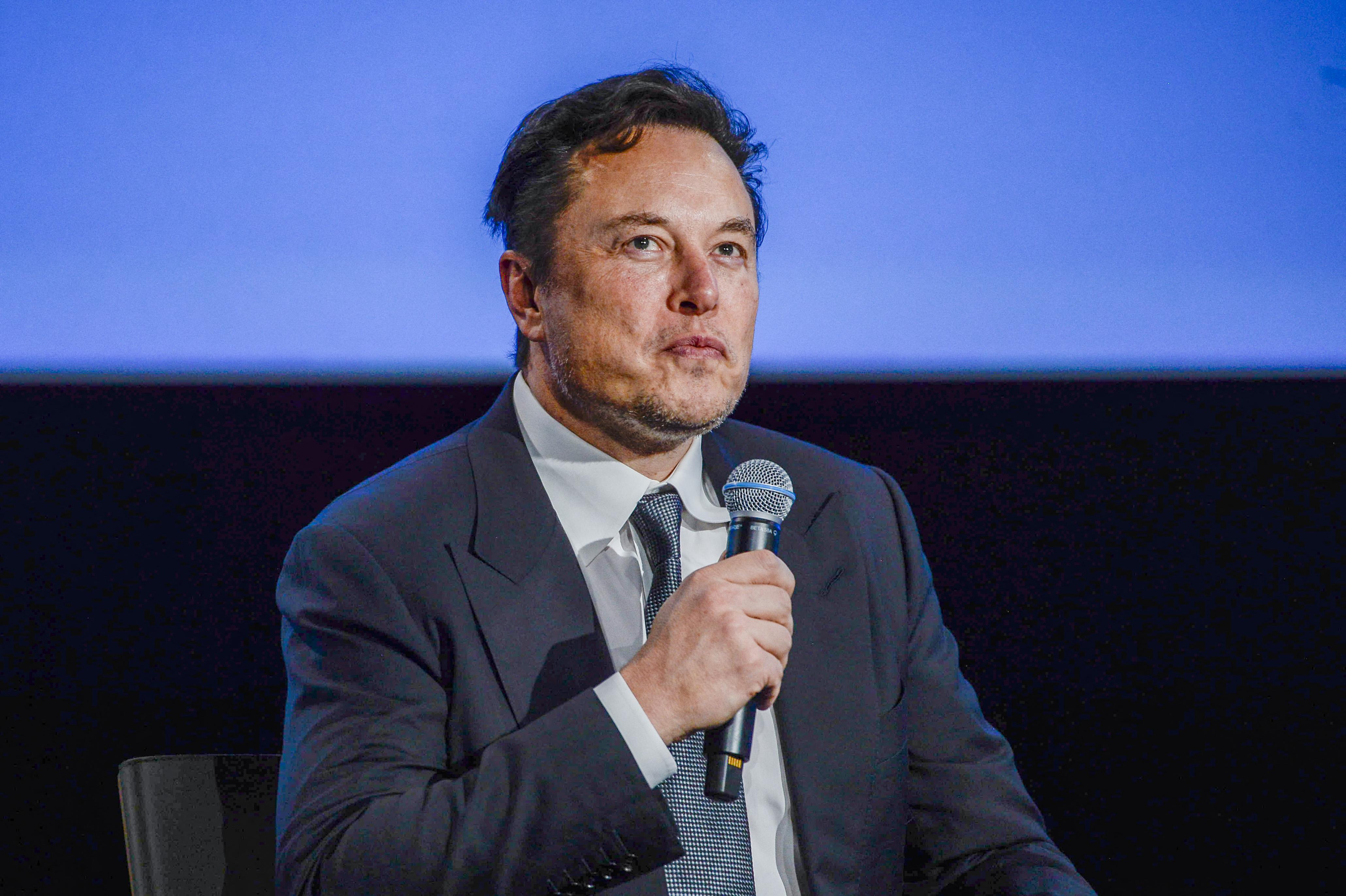Elon Musk sitting on a stage and holding a microphone