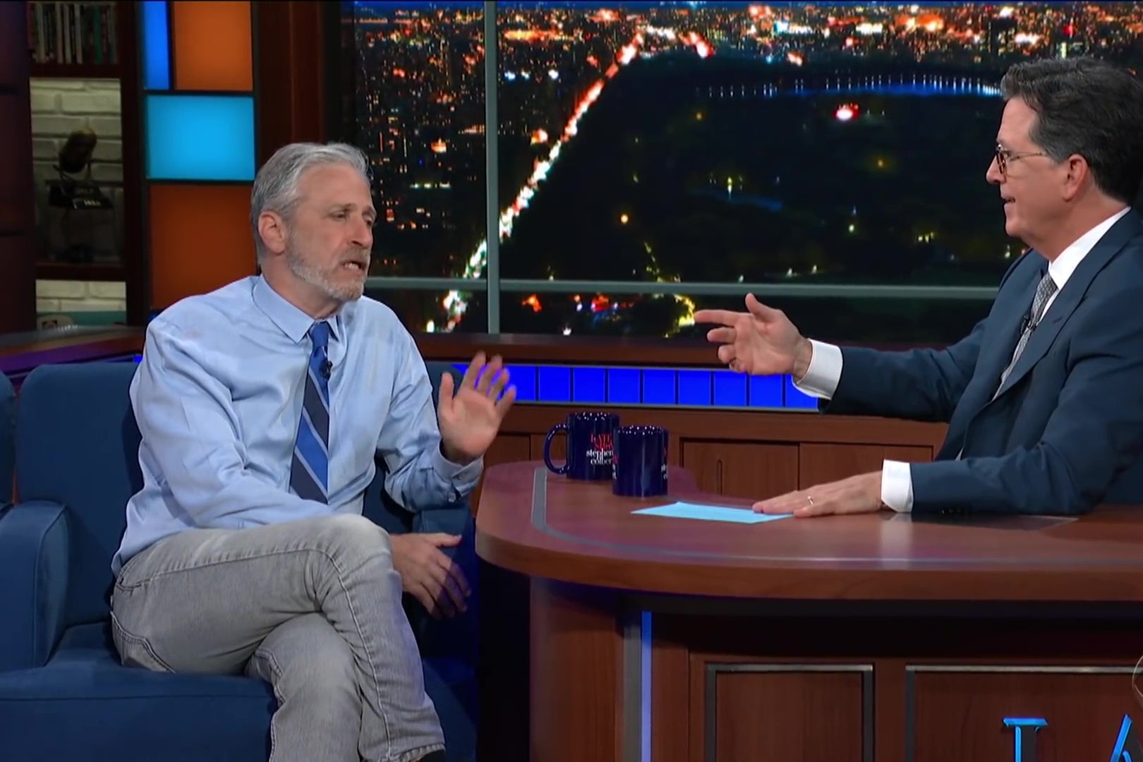 Jon Stewart, in a blue shirt, tie, and jeans, sits in the guest chair on the Late Show set, talking with Stephen Colbert, who sits behind the host's desk.