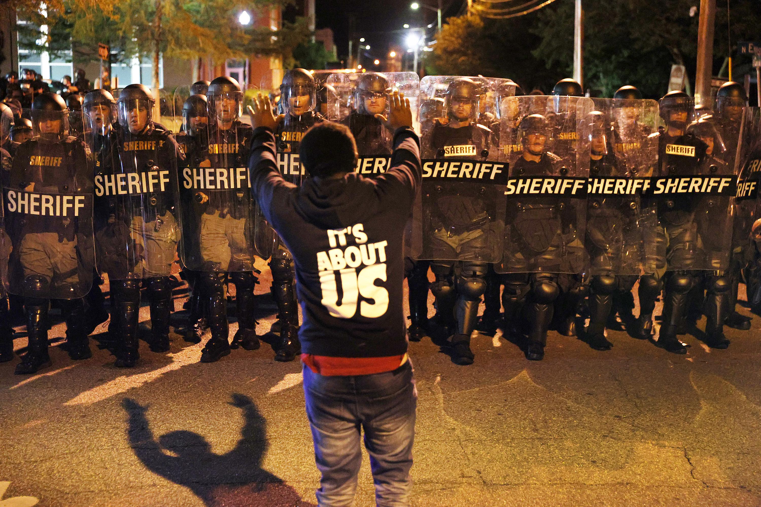 A protester stands with his arms raised in front of a line of law enforcement officials in riot gear.