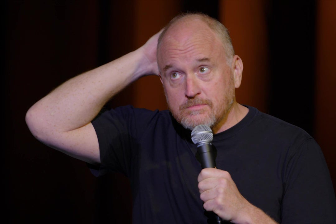 Comedian Louis C.K., wearing a blue T-shirt, holding a microphone on stage.