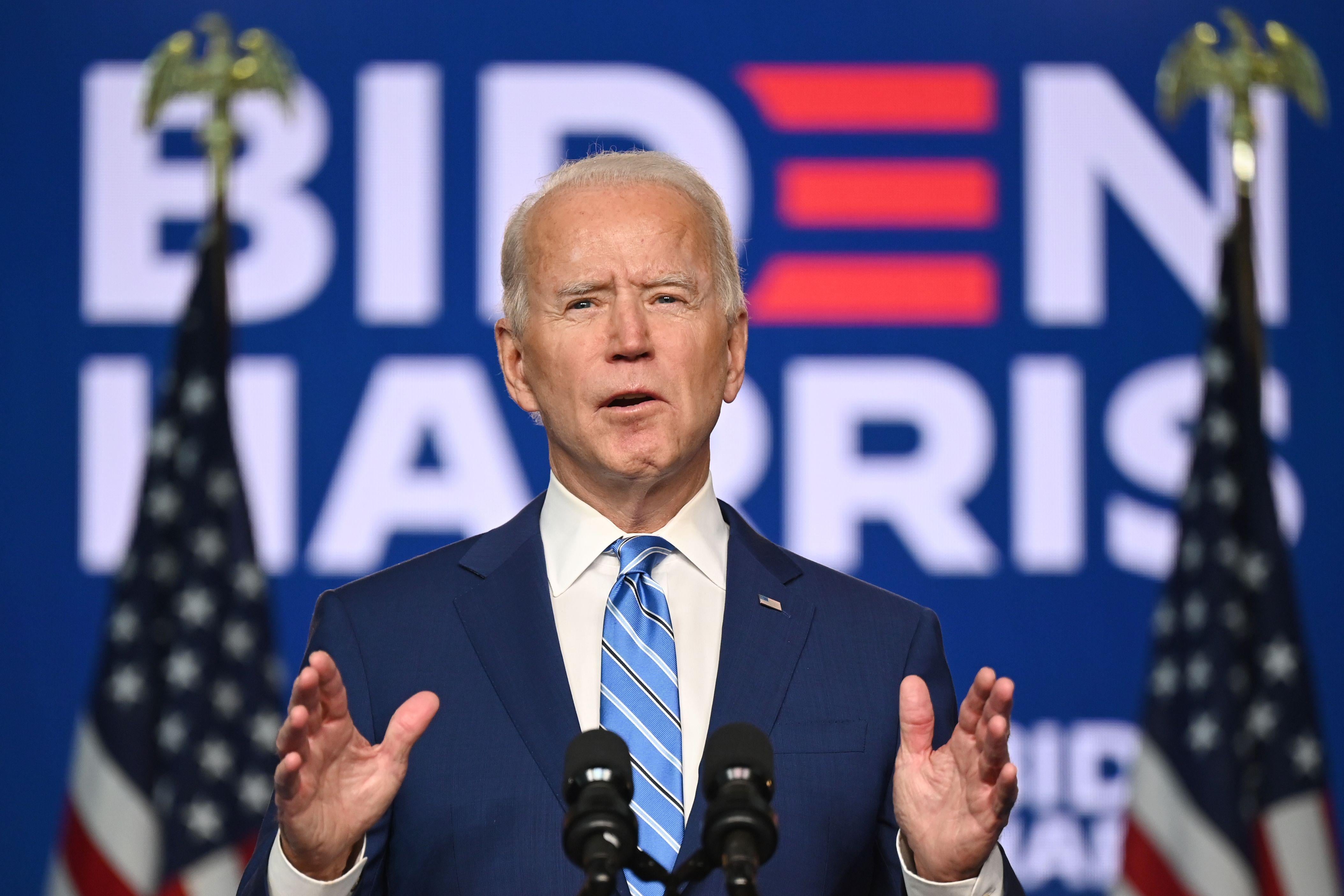 Joe Biden holds both hands up in front of him while speaking from behind a microphone.