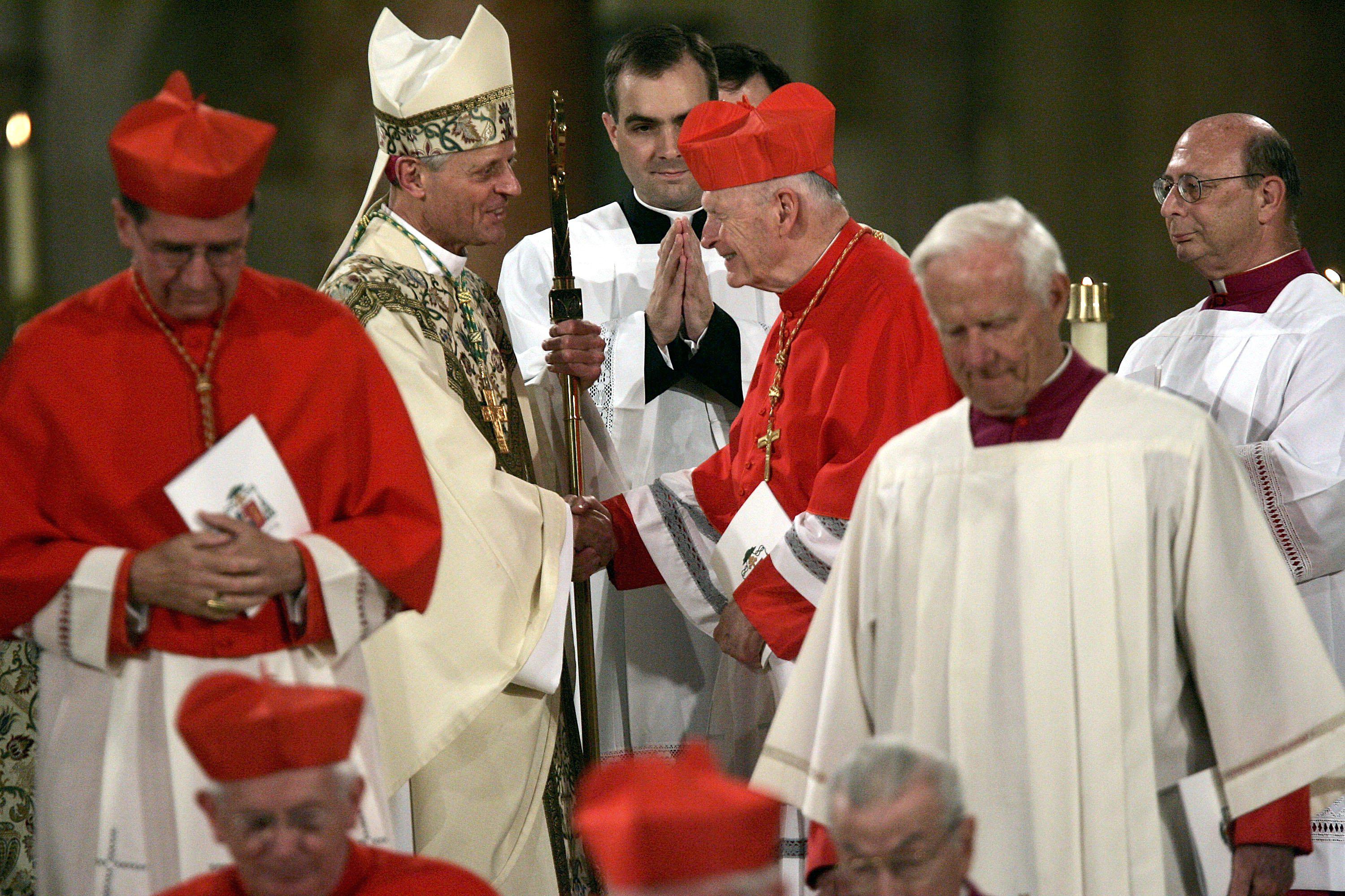Donald W. Wuerl shakes hands with disgraced Cardinal Theodore E. McCarrick (R) in the interior of a cathedral, surrounded by other members of the clergy.