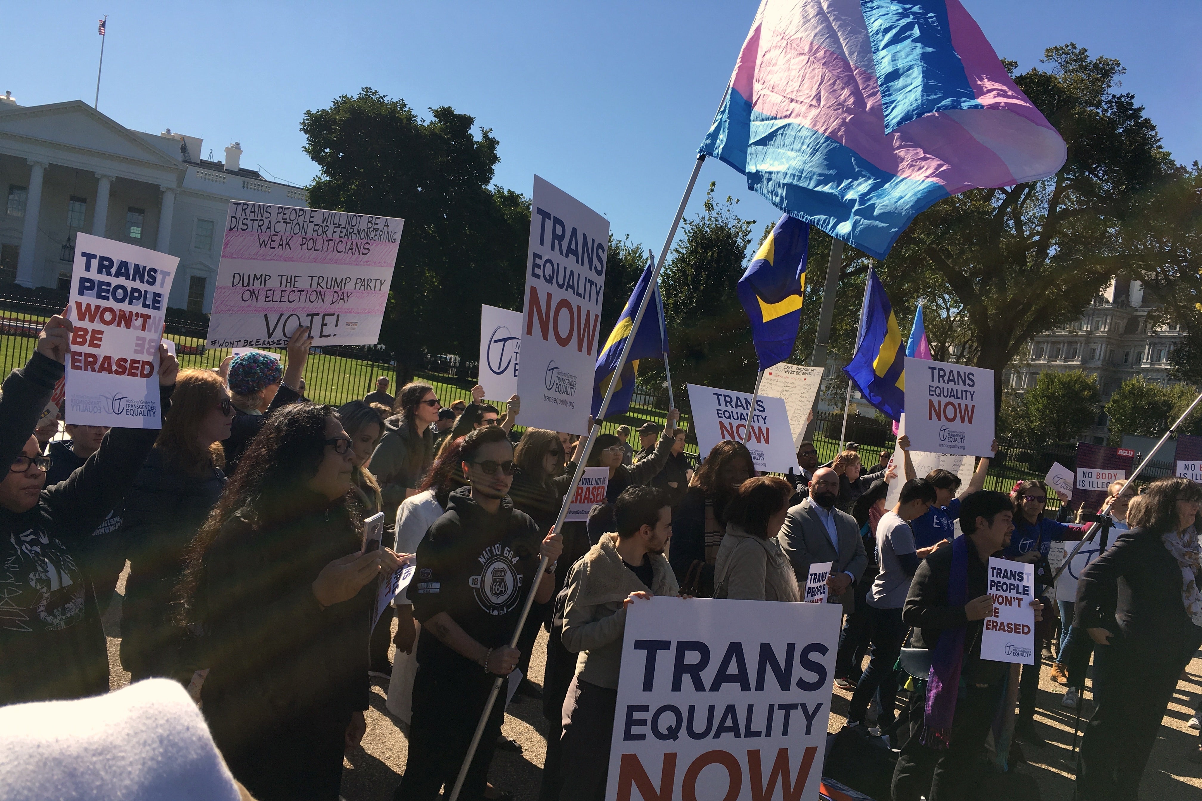 Demonstrators holding signs that say "Trans Equality Now" and "trans people won't be erased" and waving a trans flag stand in front of the White House.