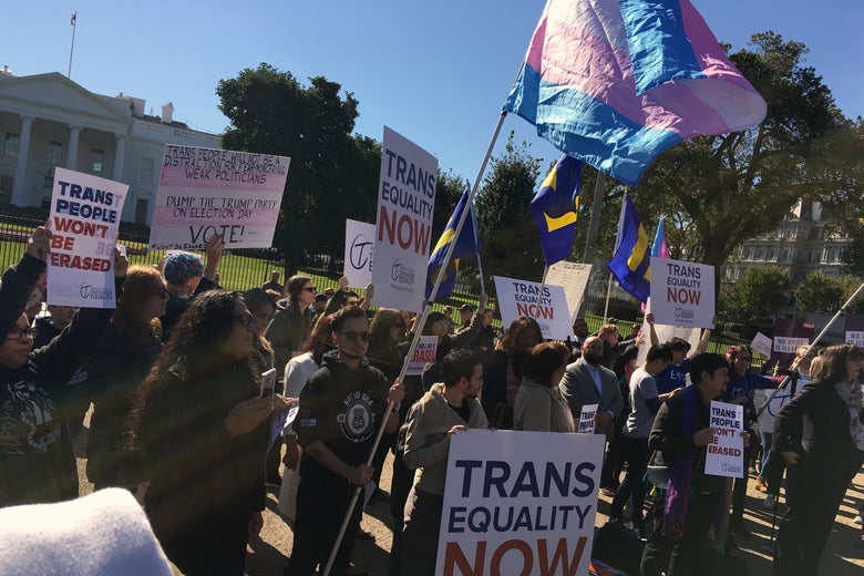 Demonstrators holding signs that say "Trans Equality Now" and "trans people won't be erased" and waving a trans flag stand in front of the White House.
