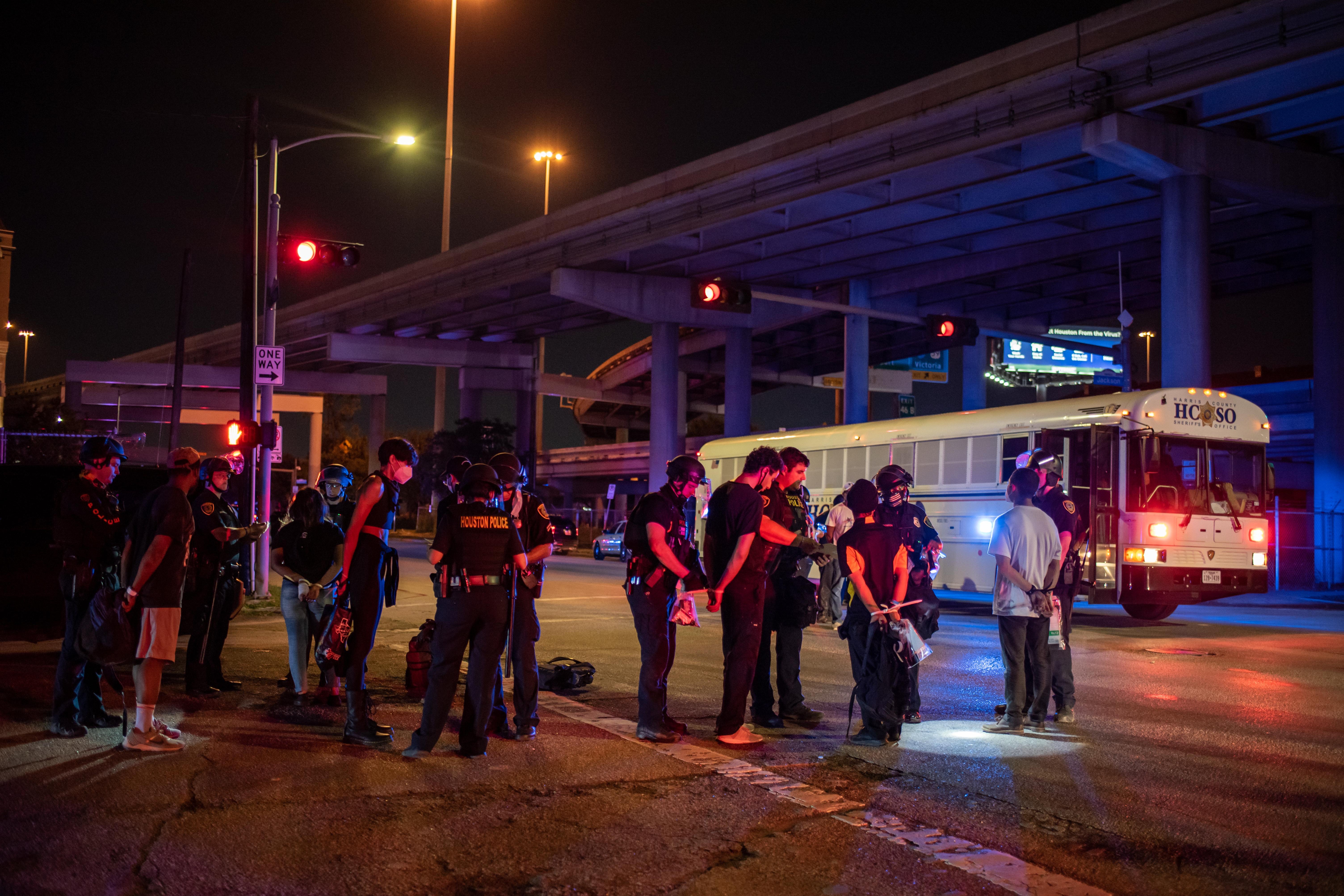 A number of handcuffed protesters with their hands tied behind their backs stand in front of a bus.