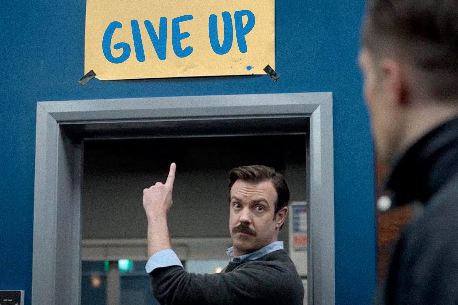 Ted Lasso pointing at the show's infamous crooked "Believe" sign changed to read "Give Up."