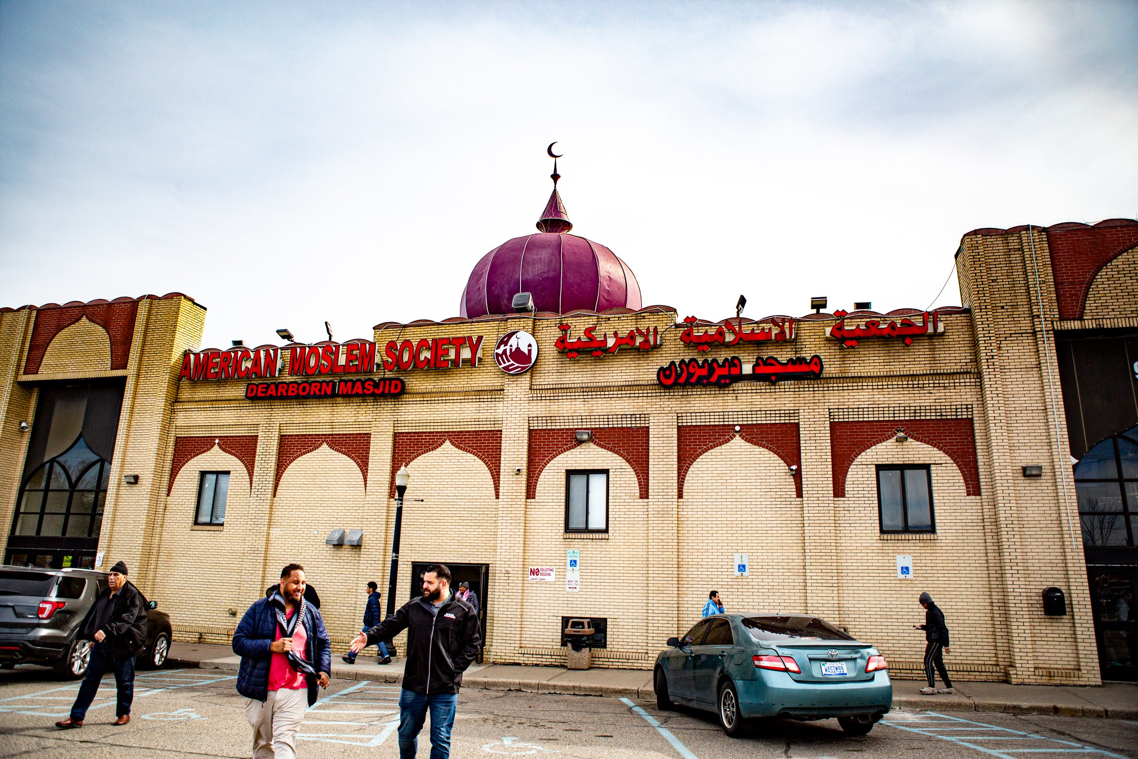 People walk into the parking lot of a brick building that says, "American Moslem Society, Dearborn Masjid" on the side.