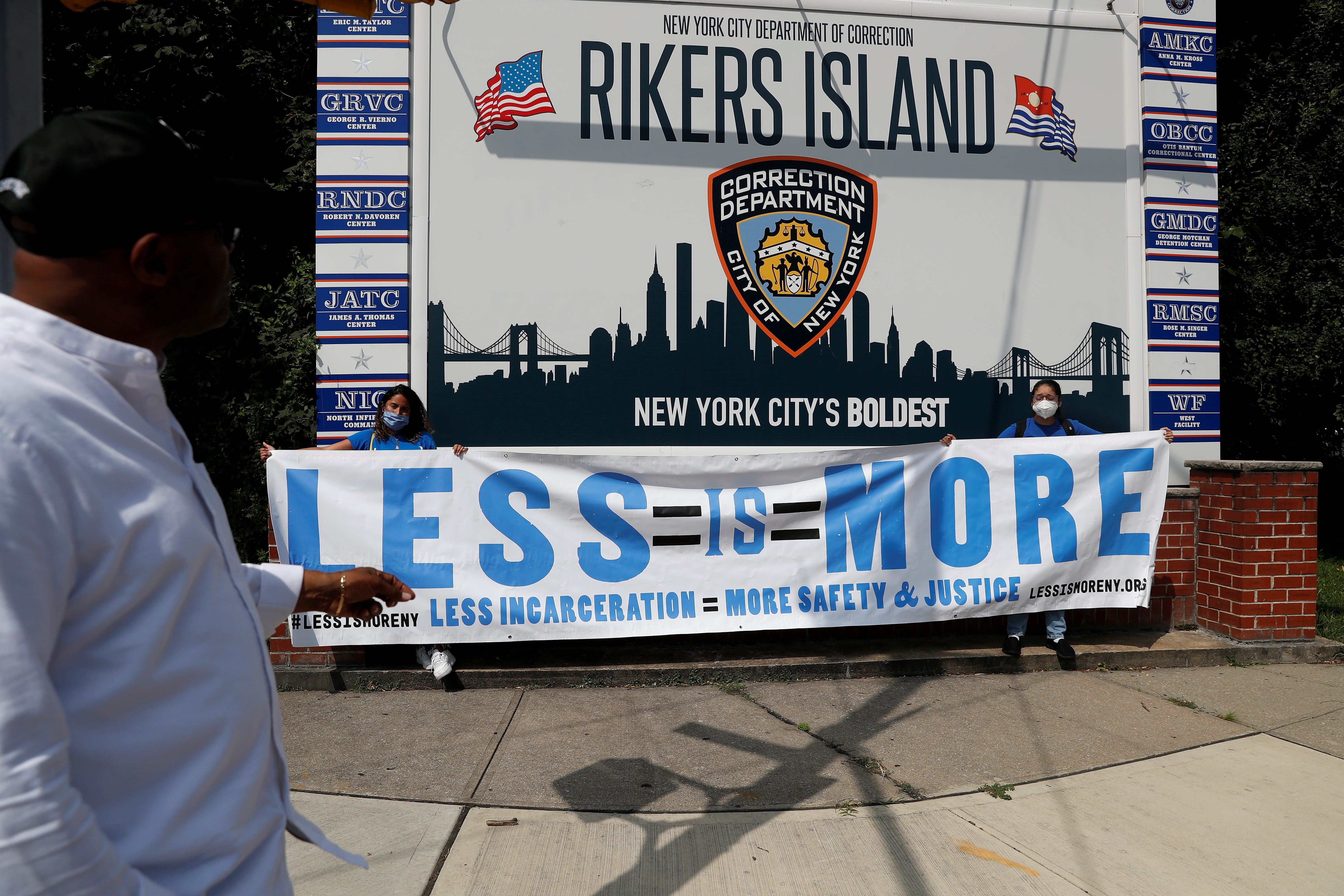 Protests hold up a large banner that says, "Less Is More" in front of the sign for Rikers Island.