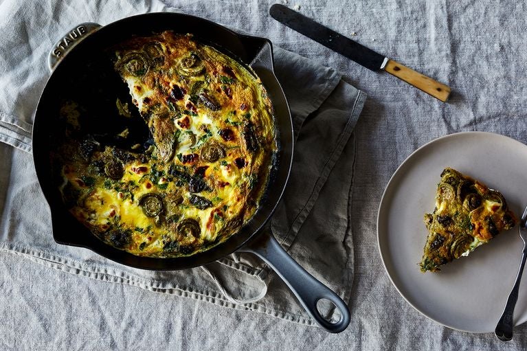 Overhead shot of cast iron pan containing a yellow fiddlehead fern frittata, with a slice carve out and sitting on a serving dish to the right of the pan.