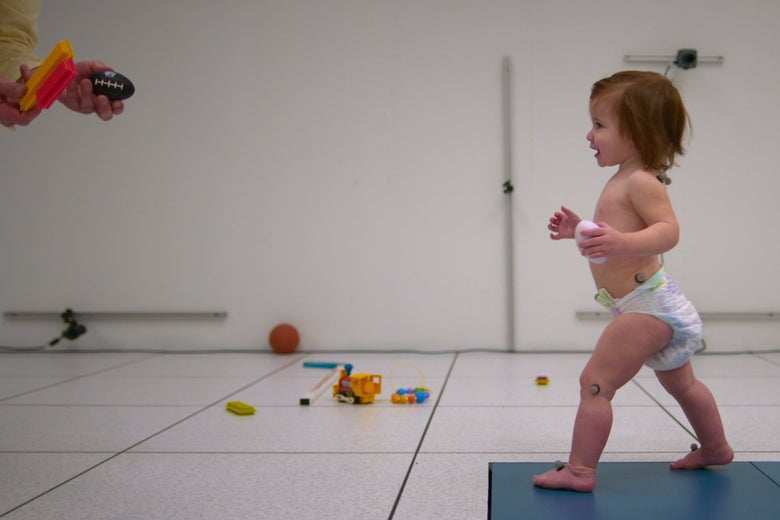 A baby in a diaper walks towards a scientist holding a toy football.