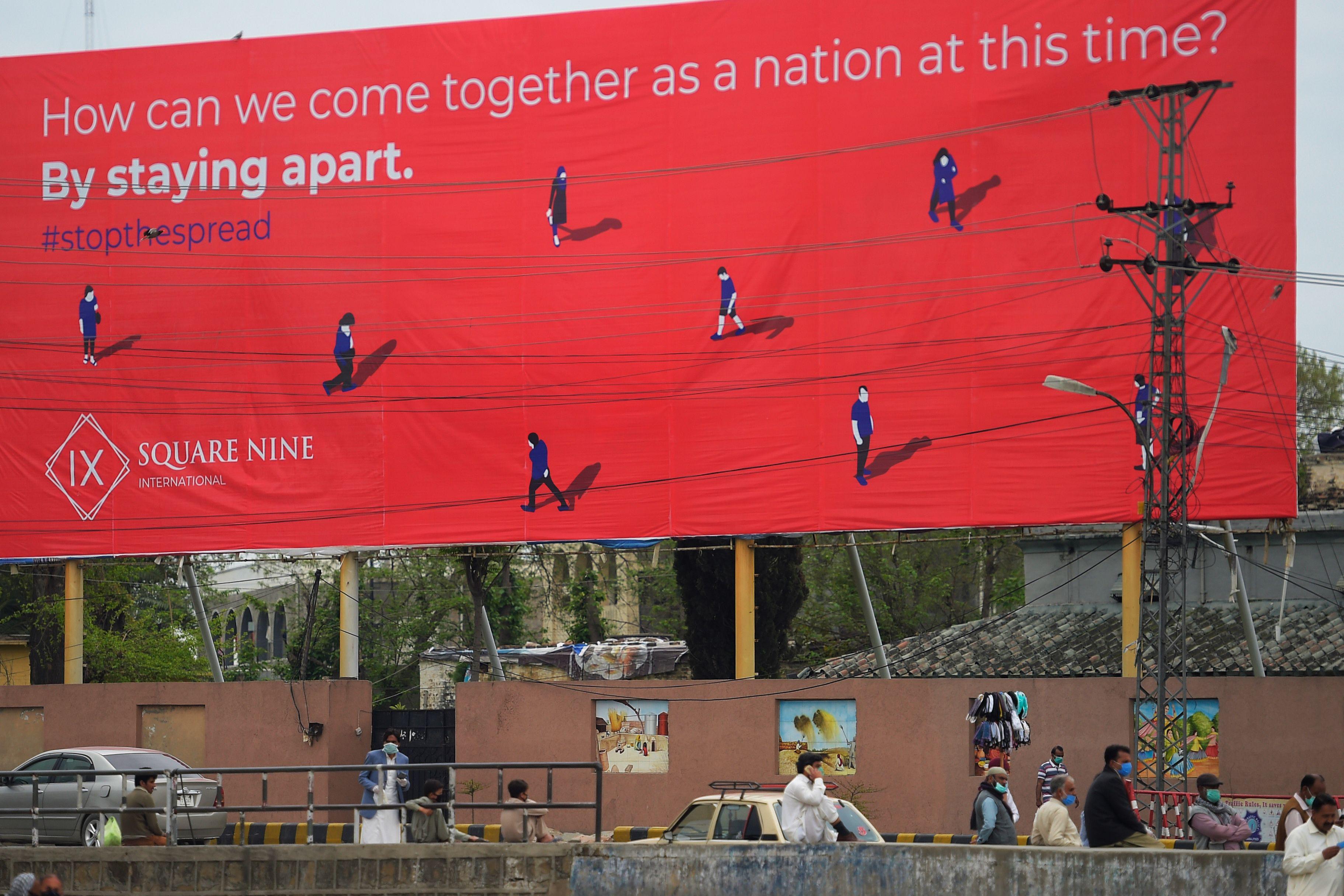 Pakistani commuters wait for transport under a billboard advocating social distancing.