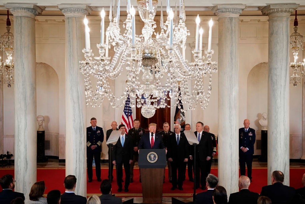 Trump is seen at the presidential lectern on a red carpet against a backdrop of white columns and a chandelier.