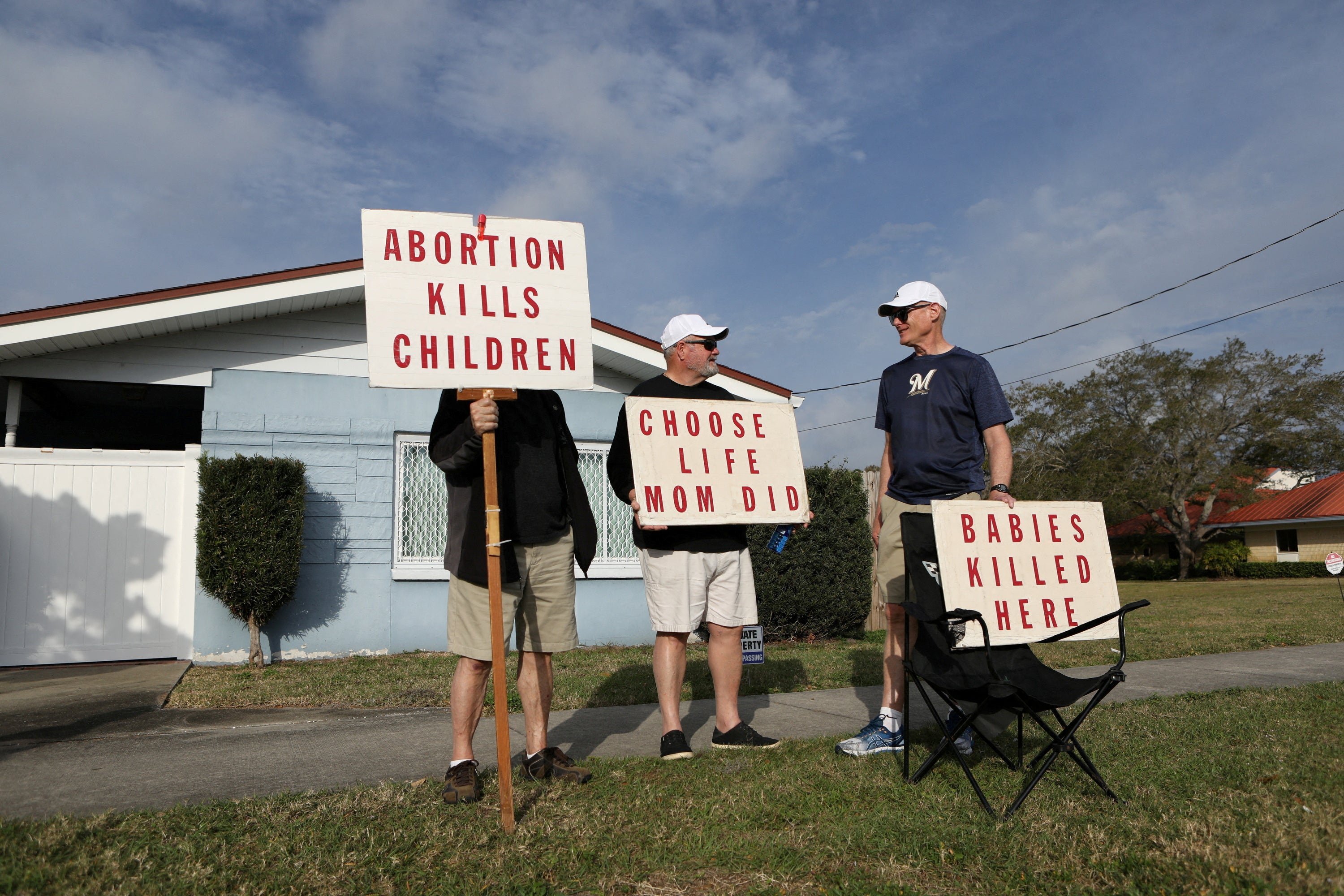 Men holding signs that say "Abortion kills children" and "Babies killed here" loiter outside of a small blue building, talking casually.