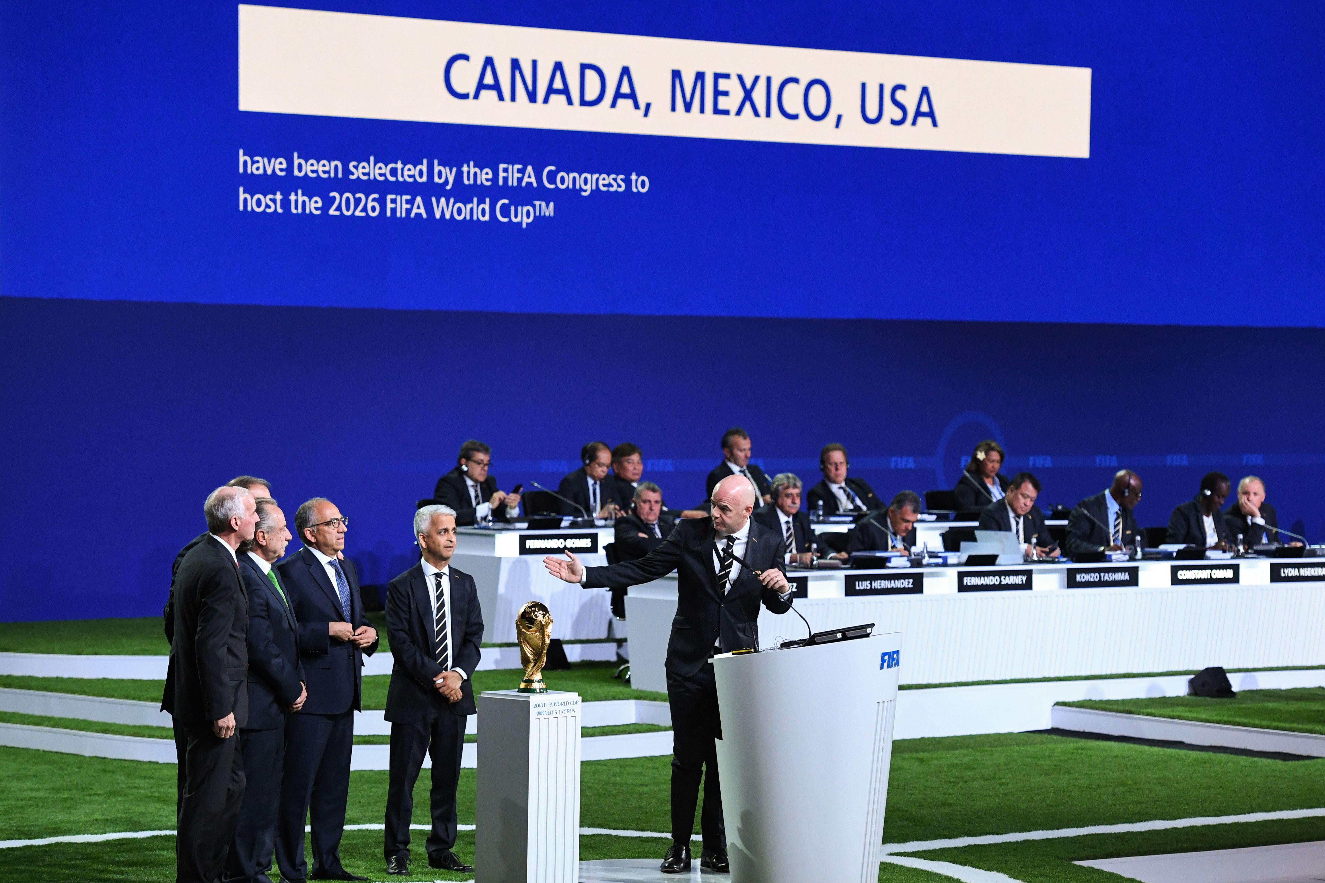 Men in suits stand to the side next to the World Cup trophy while another man in a suit talks from a white podium and gestures toward them.