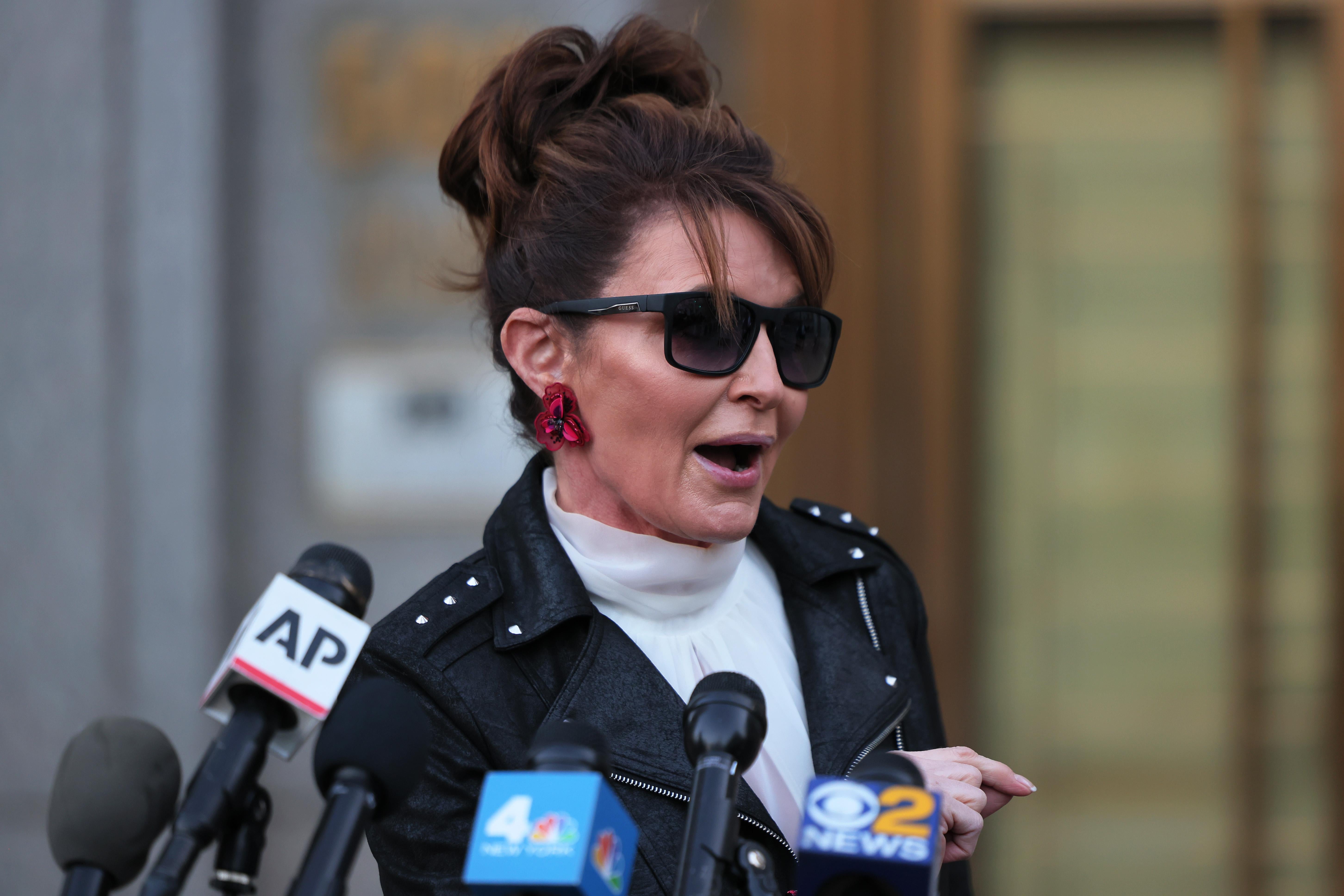 Palin in sunglasses and a black leather jacket speaks in front of a cluster of microphones outside