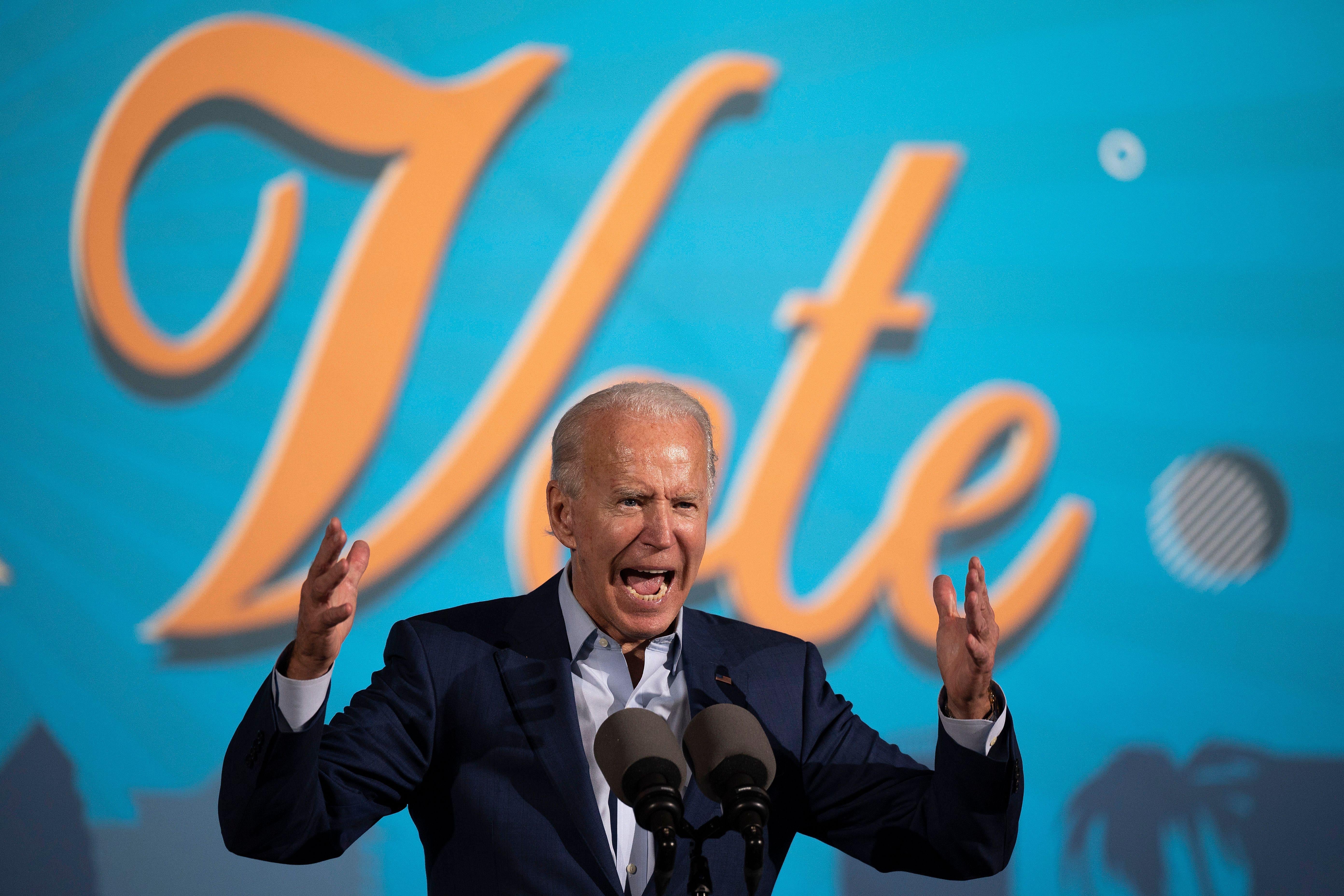 Biden stands with his arms raised, shouting at a mic. The word "vote" is on a backdrop behind him.