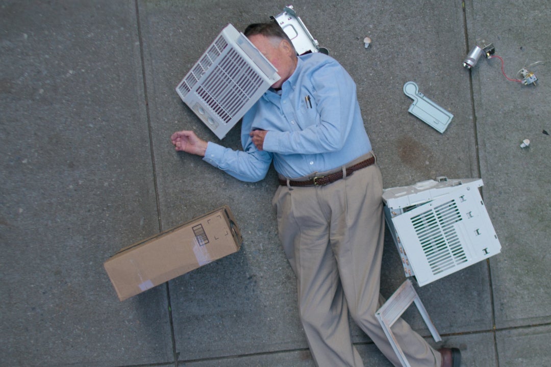 An older white man lies sprawled on the pavement, in loose khakis and a blue Oxford tucked into his waist, the pieces of a broken AC unit scattered around him