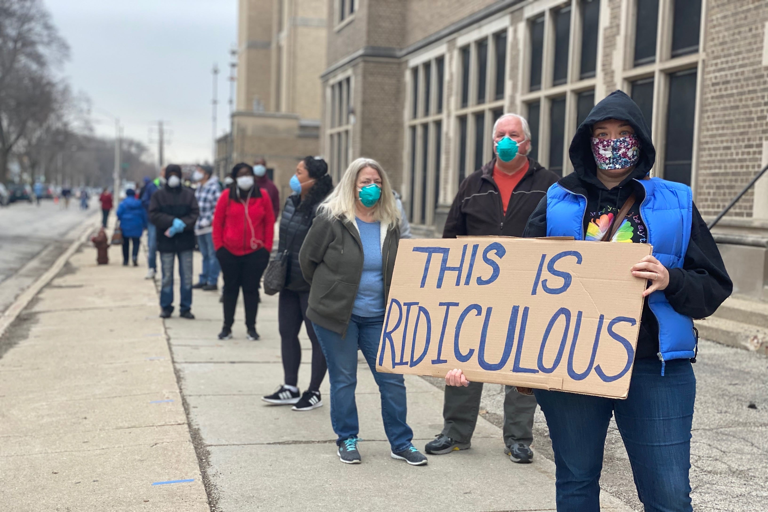 A line of voters spread out on a sidewalk and wearing masks. In the front, a masked woman holds a sign that says "This Is Ridiculous."