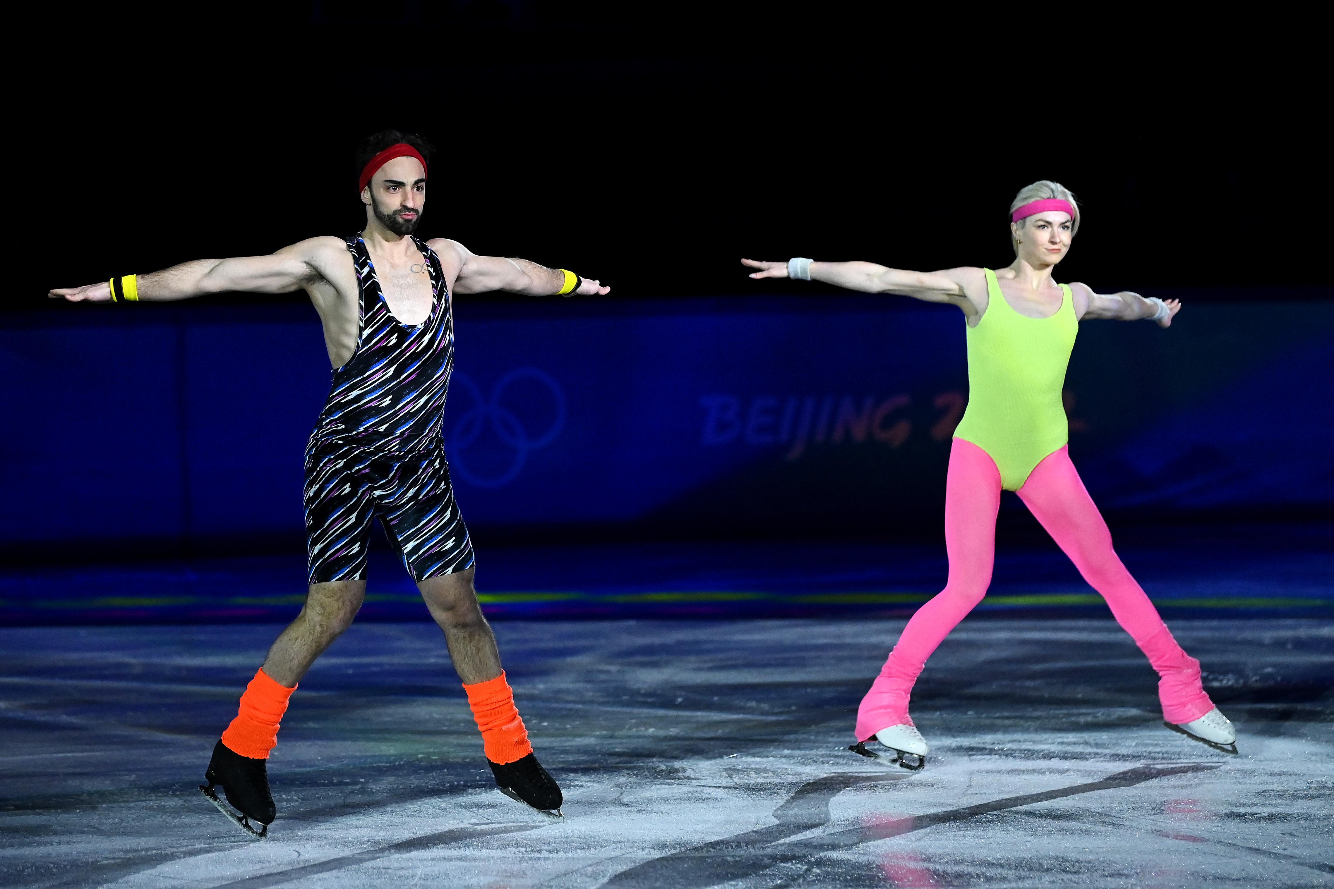 The couple in 80s fitness outfits, man in leotard woman in neon leggings and top, both in ankle warmers and headbands, with their arms outstretched in unison to the side