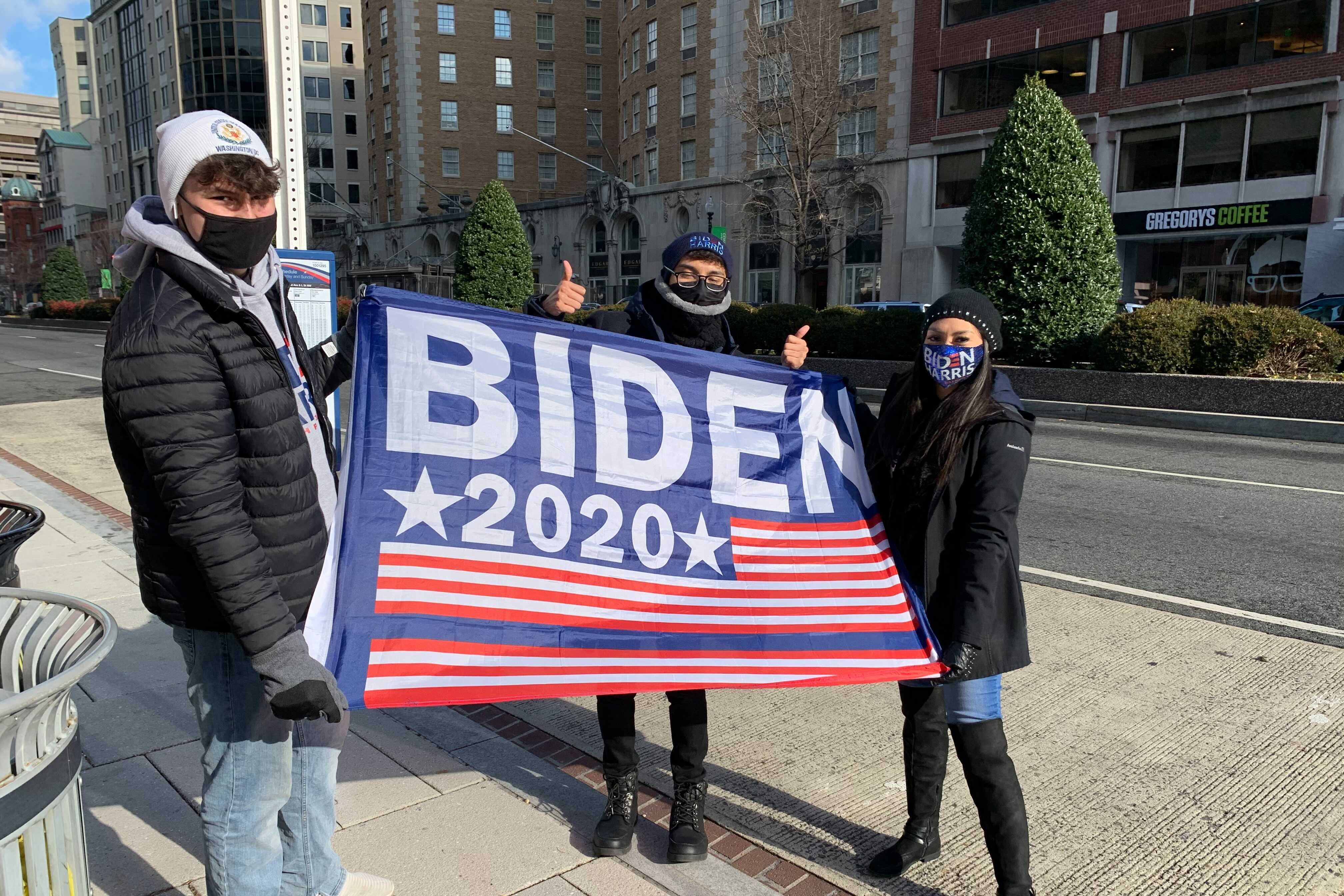 Balian Sanders, Bryan Godinez, and Yessica Leon stand on a sidewalk holding a Biden 2020 flag.