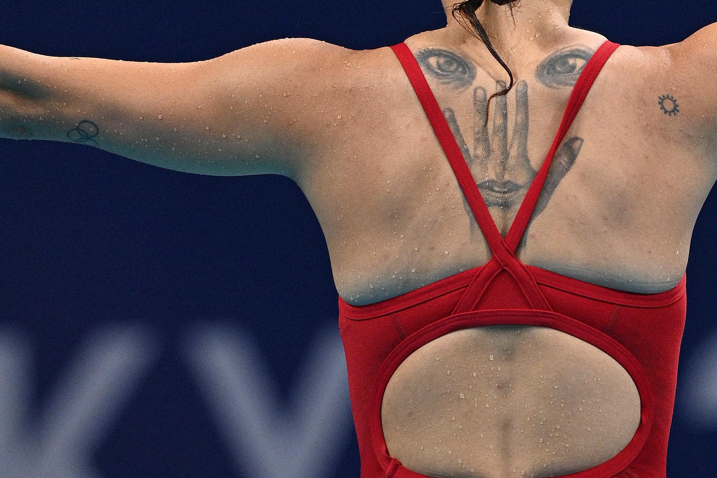 The back of a female diver in her competitive bathing suit, stretching her arms out to the sides, a tattoo visible between her shoulder blades of two eyes, below which are inked the back of a hand and a mouth inside the base of that hand