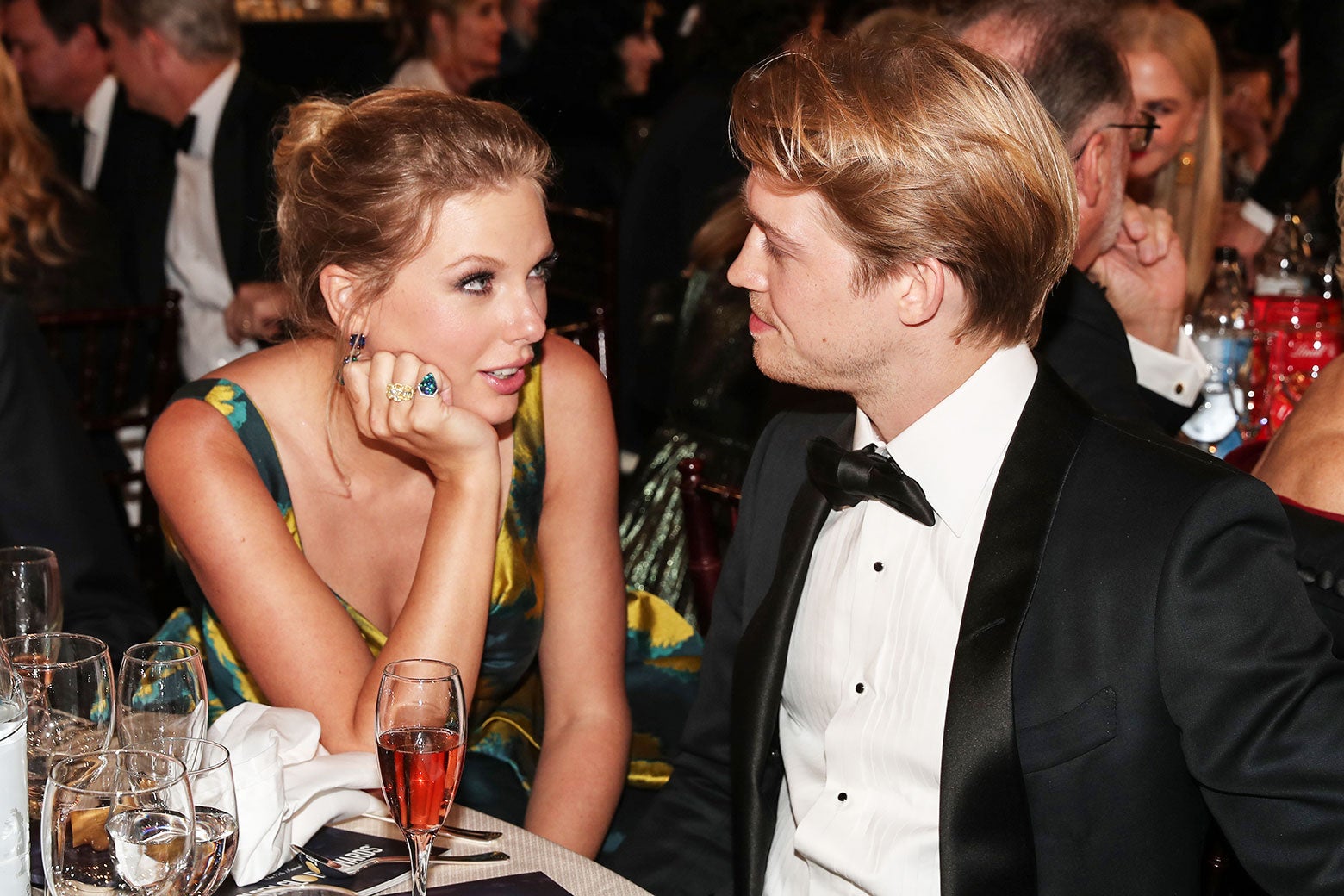 Taylor and Joe sit next to each other at a dinning table in formal wear. Taylor rests her chin on her hand and looks into Joe's eyes.