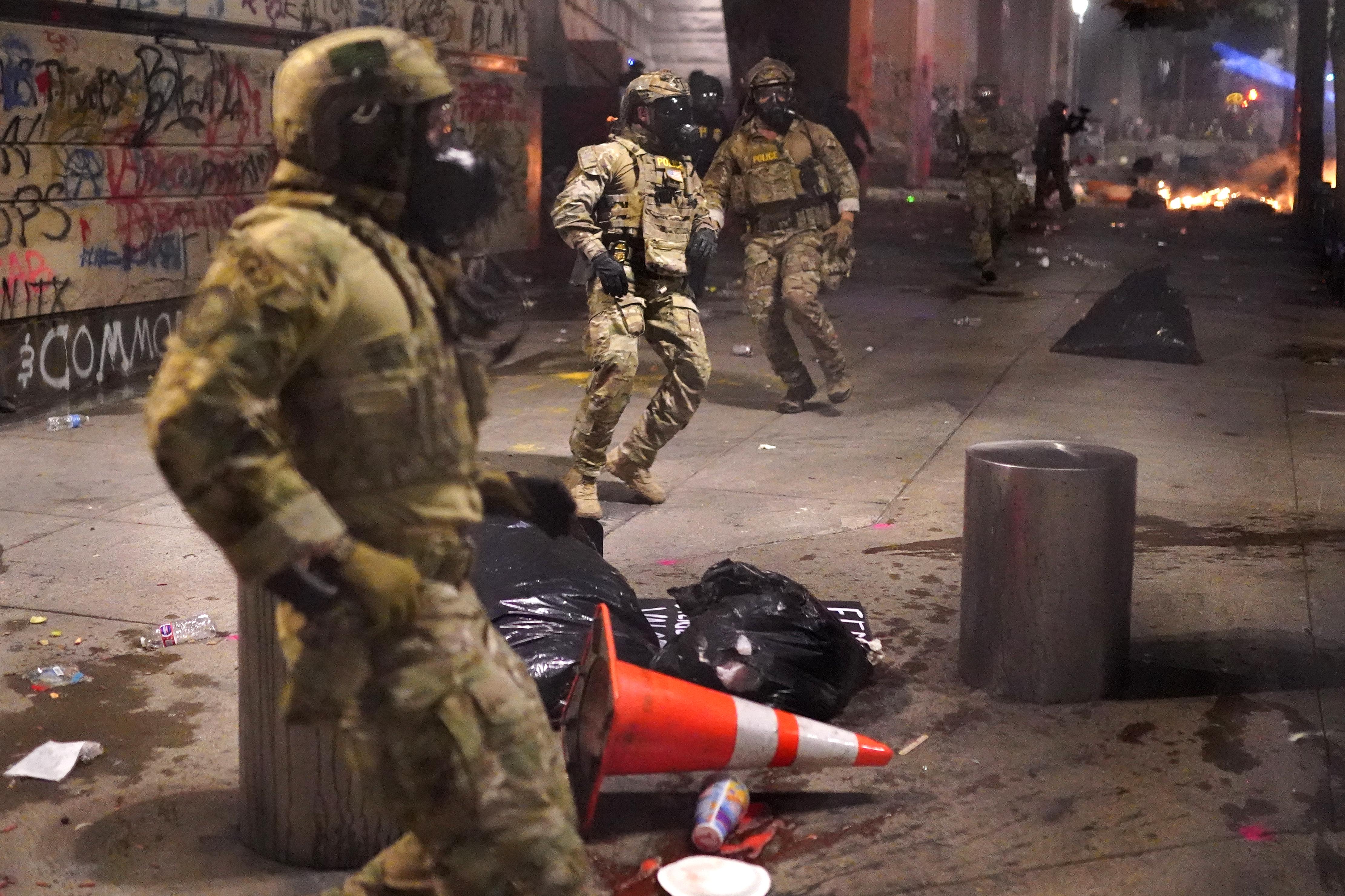 Federal officers in gas masks and camo gear surrounded by trash and graffiti.