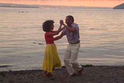 A man and woman dance on the shore of a lake.