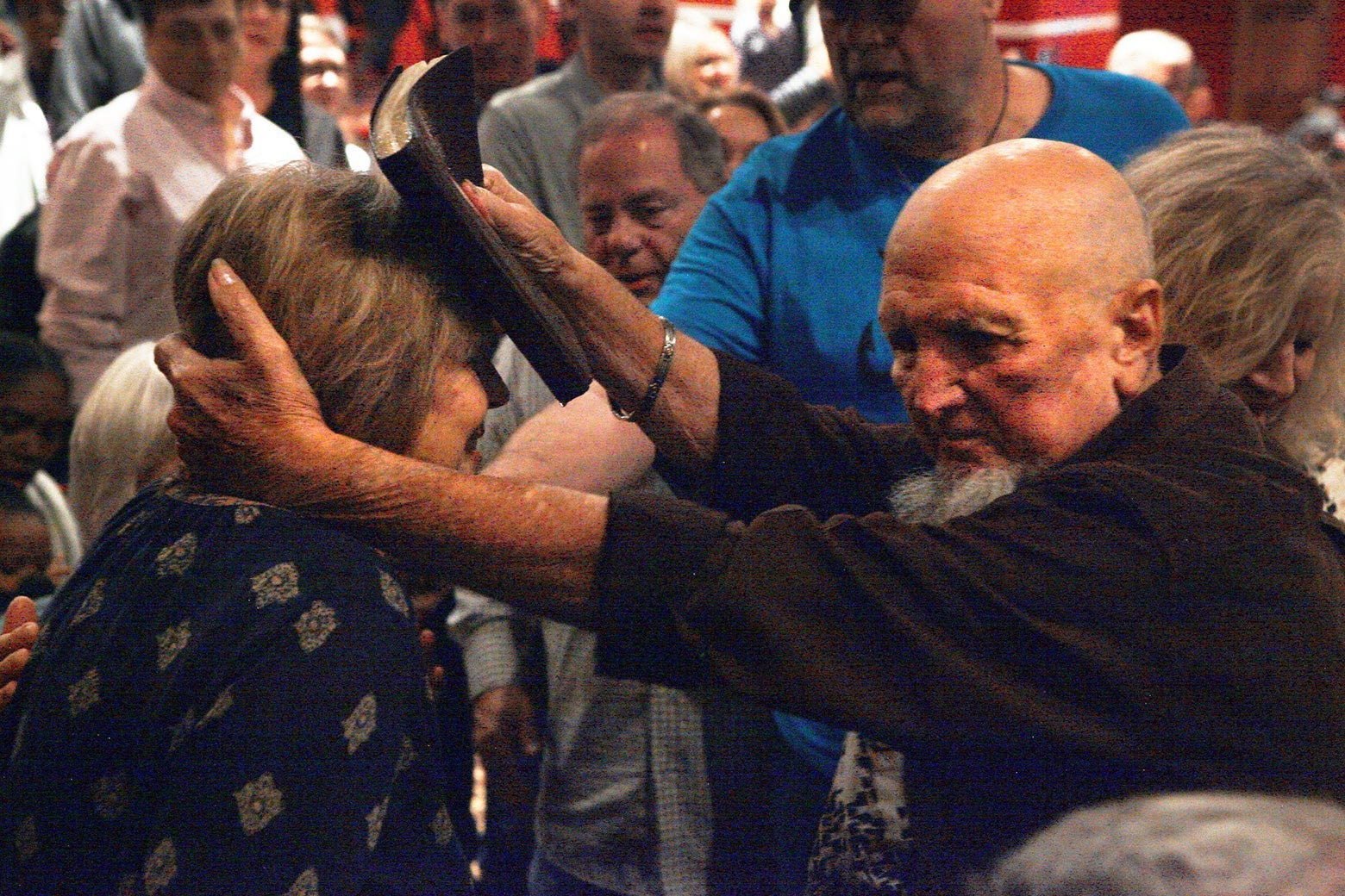 A bearded man presses an oily Bible to a woman's head as people surrounding them watch.