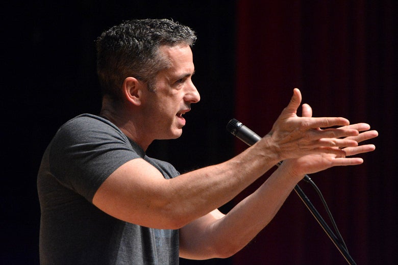 Dan Savage, seen in profile, holds his hands out in front of him as he speaks into a microphone.