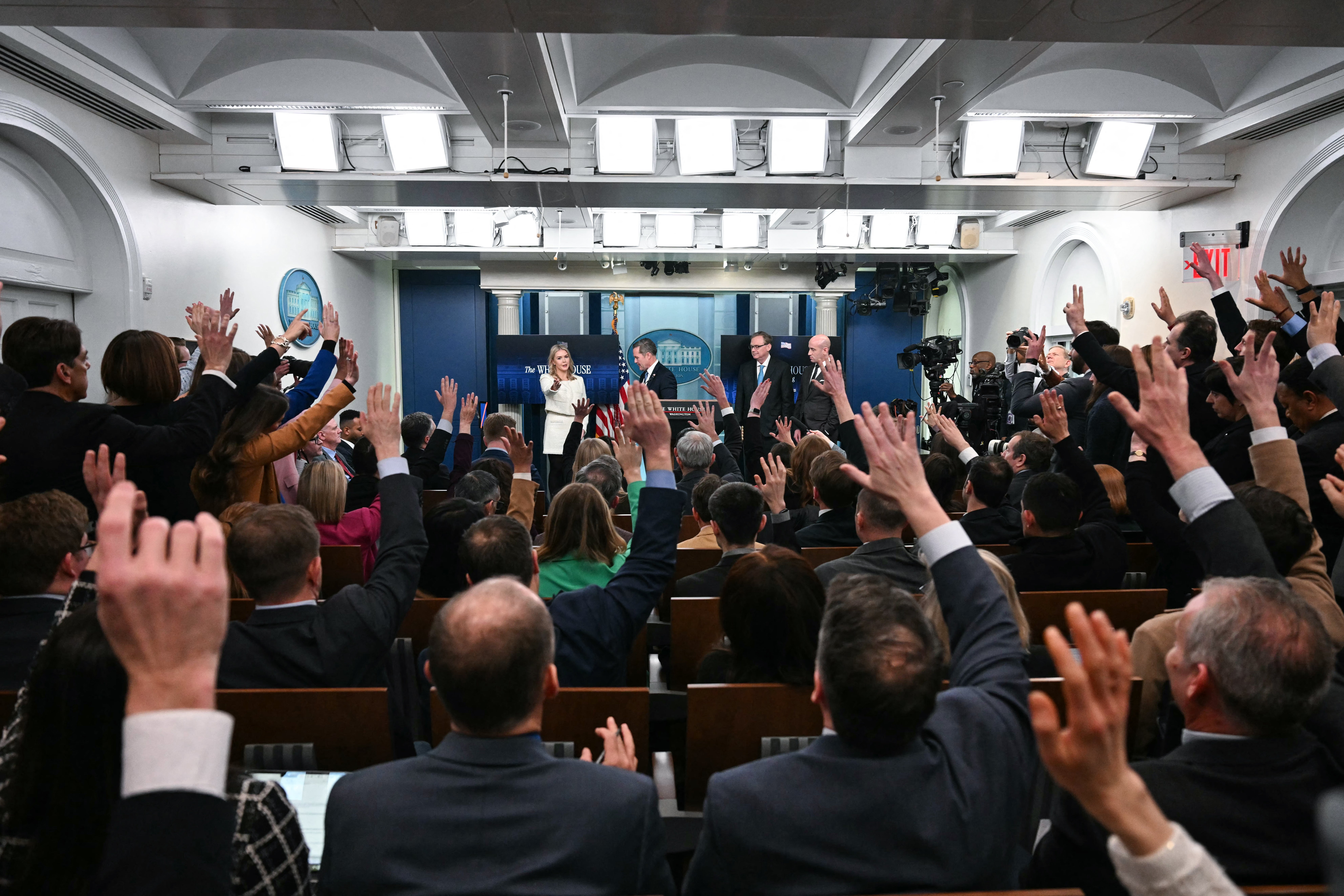 In the foreground, crowds of reporters seated and standing have their hands raised. In the background, White House Press Secretary Karoline Leavitt, US National Security Advisor Mike Waltz, Director of the National Economic Council Kevin Hassett and White House deputy chief of staff for policy/homeland security advisor Stephen Miller take questions during the daily press briefing in the Brady Briefing Room of the White House.