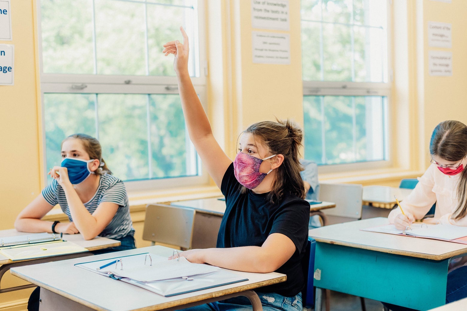 Three students wearing masks sit at spaced-out desks in a classroom. One student raises her hand.