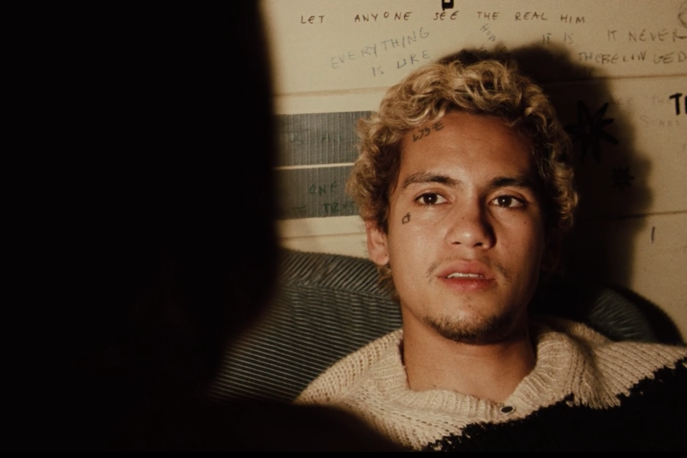 A boy with a face tattoo and blonde hair sits in front of a wall with graffiti. 