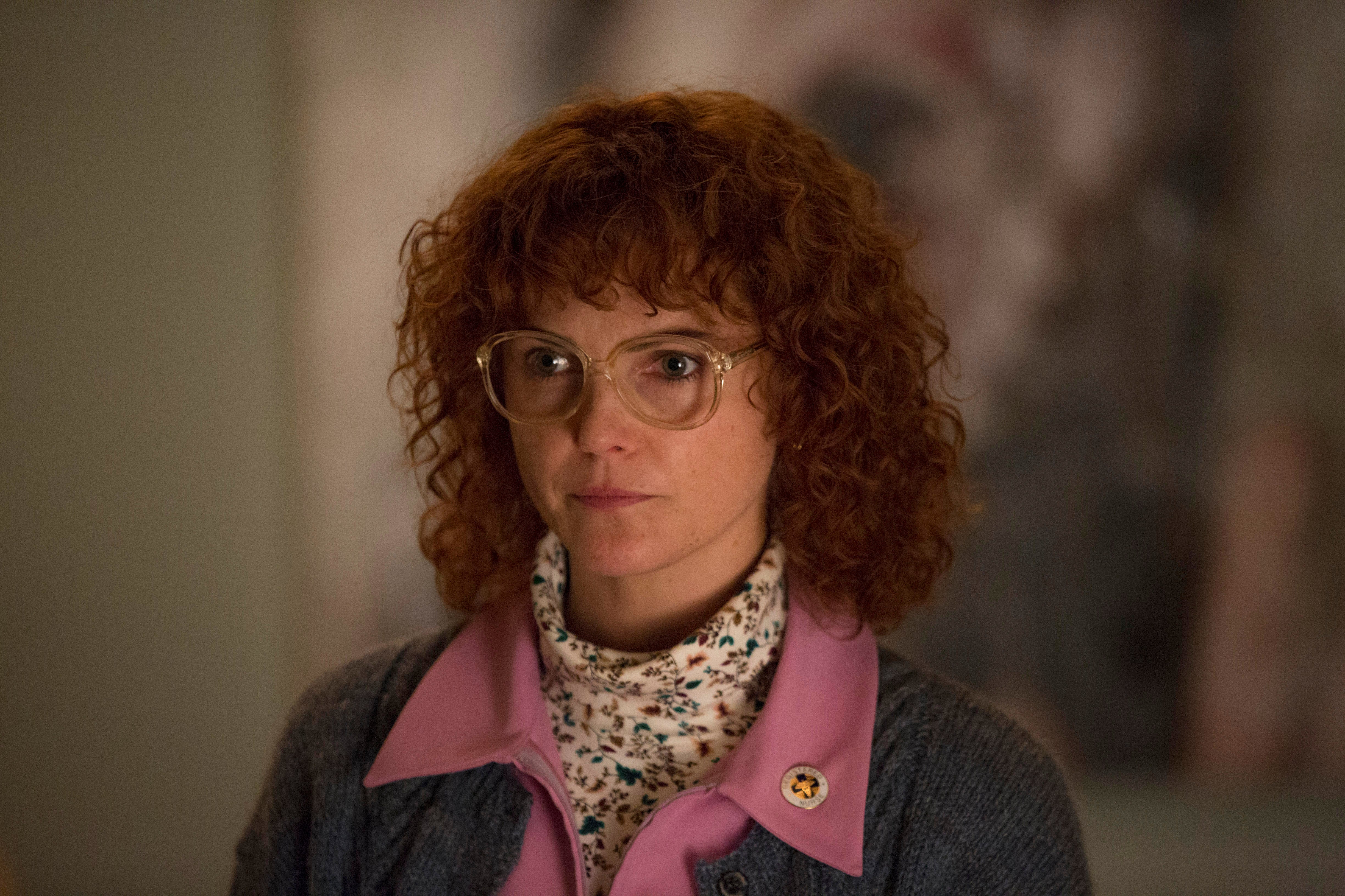 Keri Russell plays Elizabeth in The Americans. In this still, Elizabeth is in disguise as Stephanie, a dowdy home care nurse. She wears big, round glasses, a crazy-curly redhead wig, and a speckle-patterned white fluffy turtleneck under a pink-collared denim shirt.