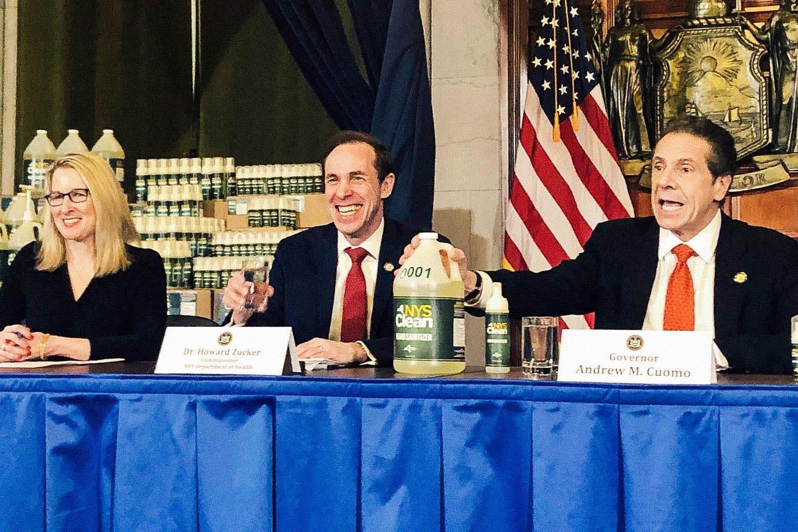 Cuomo, seated beside Dr. Howard Zucker on a panel, presents a jug of hand sanitizer at a briefing.
