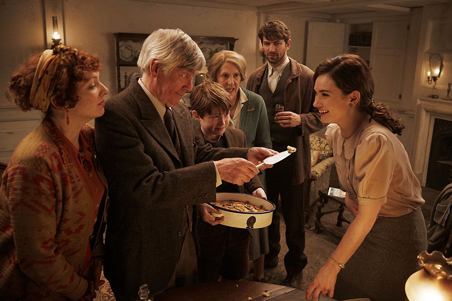 In a living room, Kit Connor holds a dish and Tom Courtenay offers Lily James a bite of food. They are standing in a living room with Michiel Huisman, Penelope Wilton, and Katherine Parkinson.