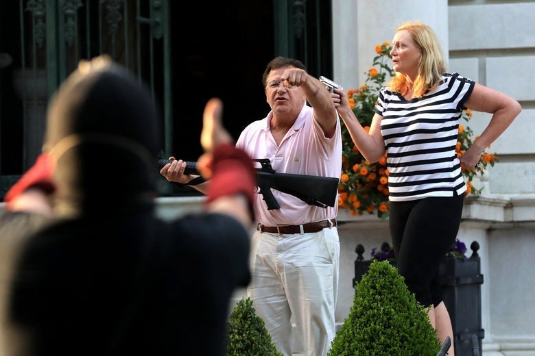Mark, wearing a pink polo and khakis and holding a semi-automatic rifle, stands in front of his house and yells at a protester in the foreground. Patricia stands beside him, wearing a striped shirt and capris and pointing a handgun to her right.