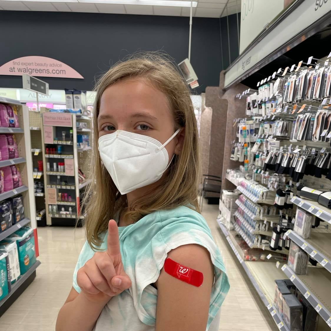 A girl standing in a pharmacy holds her finger up. She is wearing a mask and a rolled up sleeve shows a band-aid on her arm.