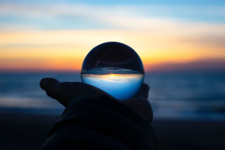 A hand holds a crystal ball in front of a beach sunrise or sunset.