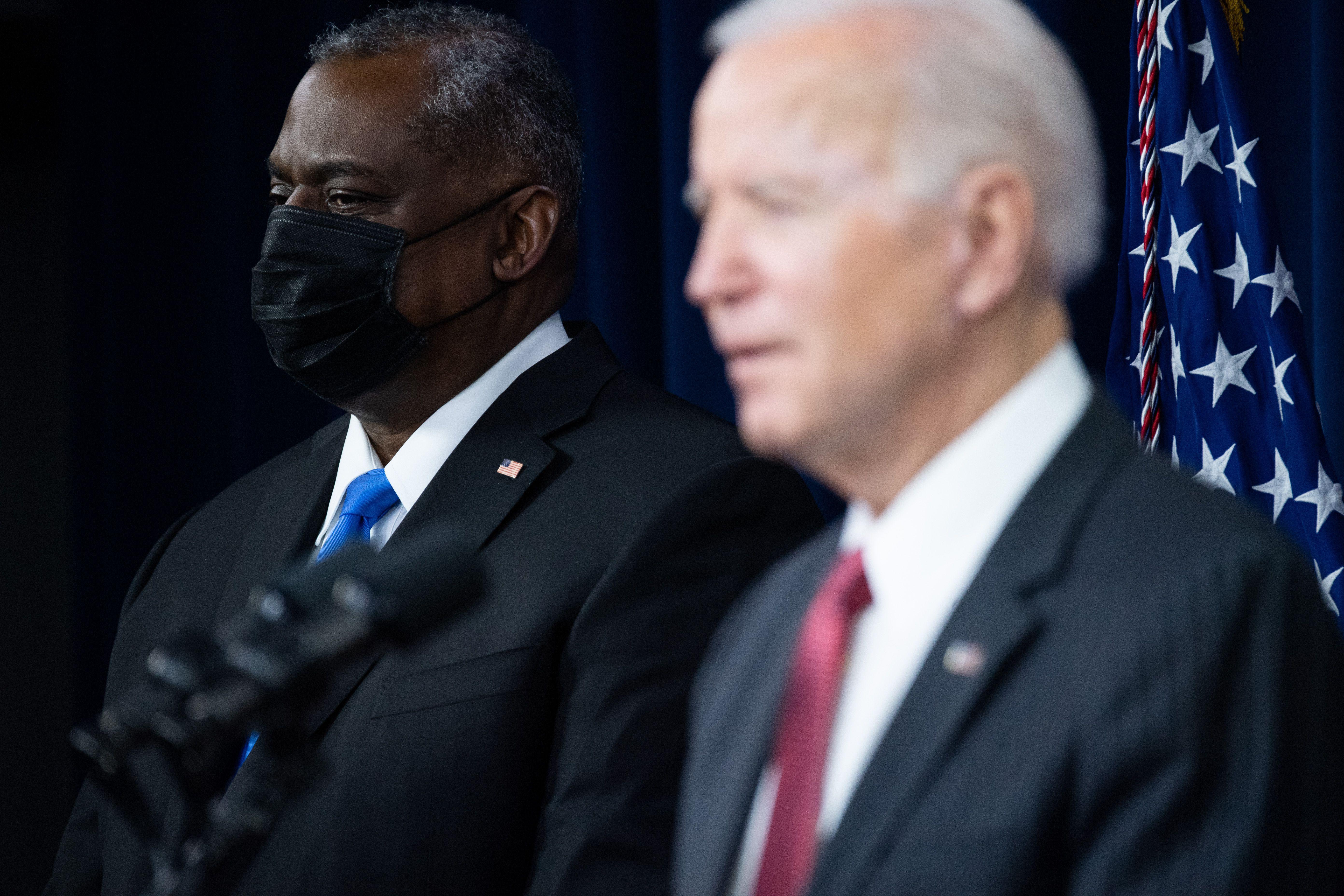 Biden speaks in the foreground and Secretary of Defense Lloyd Austin, wearing a black mask, stands in the background during a press conference at the Pentagon.