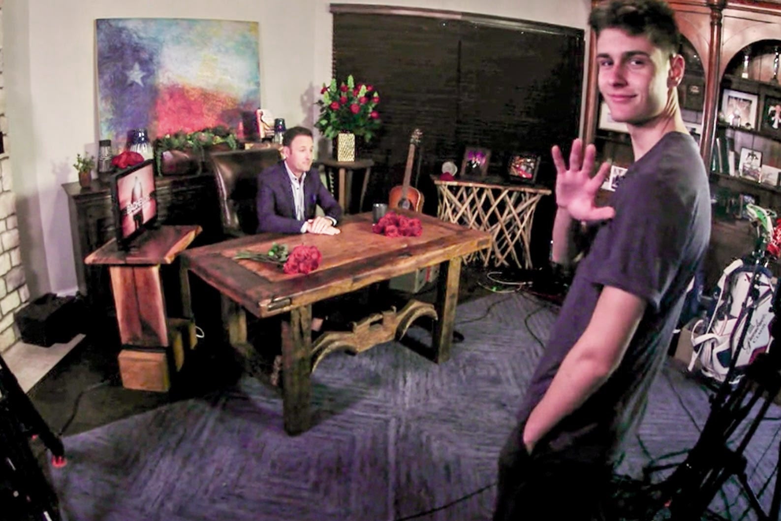 Joshua waving in the foreground as his father sits at a desk in the background in a makeshift set.