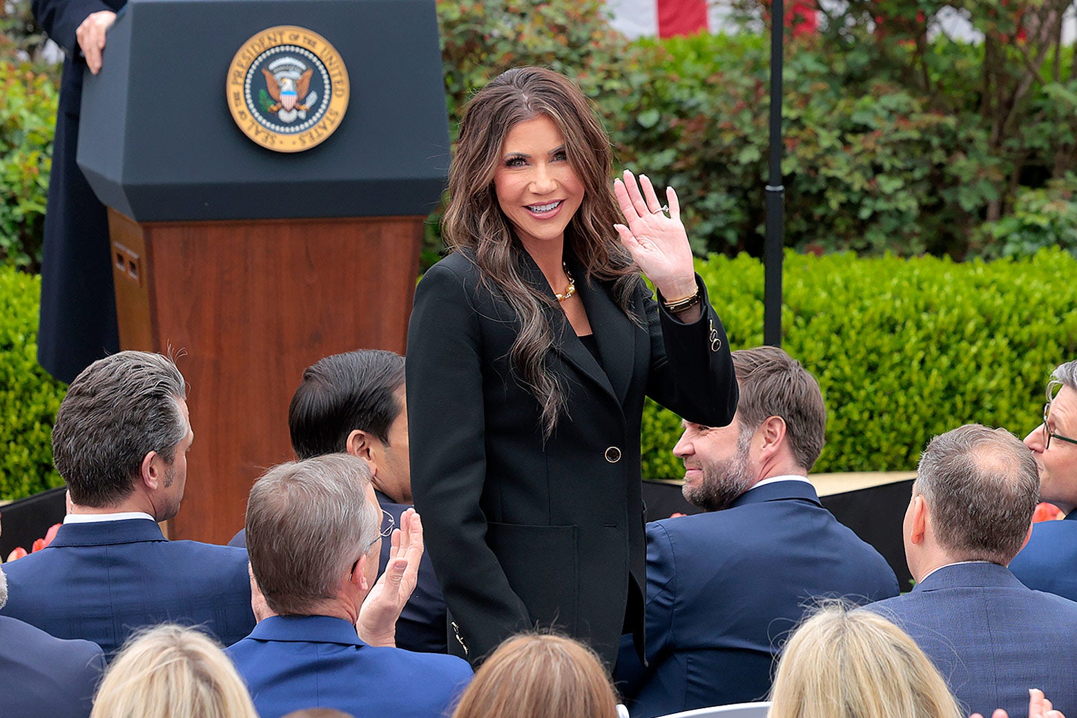 Kristi Noem standing up in a crowd of men in blue suits and waving.