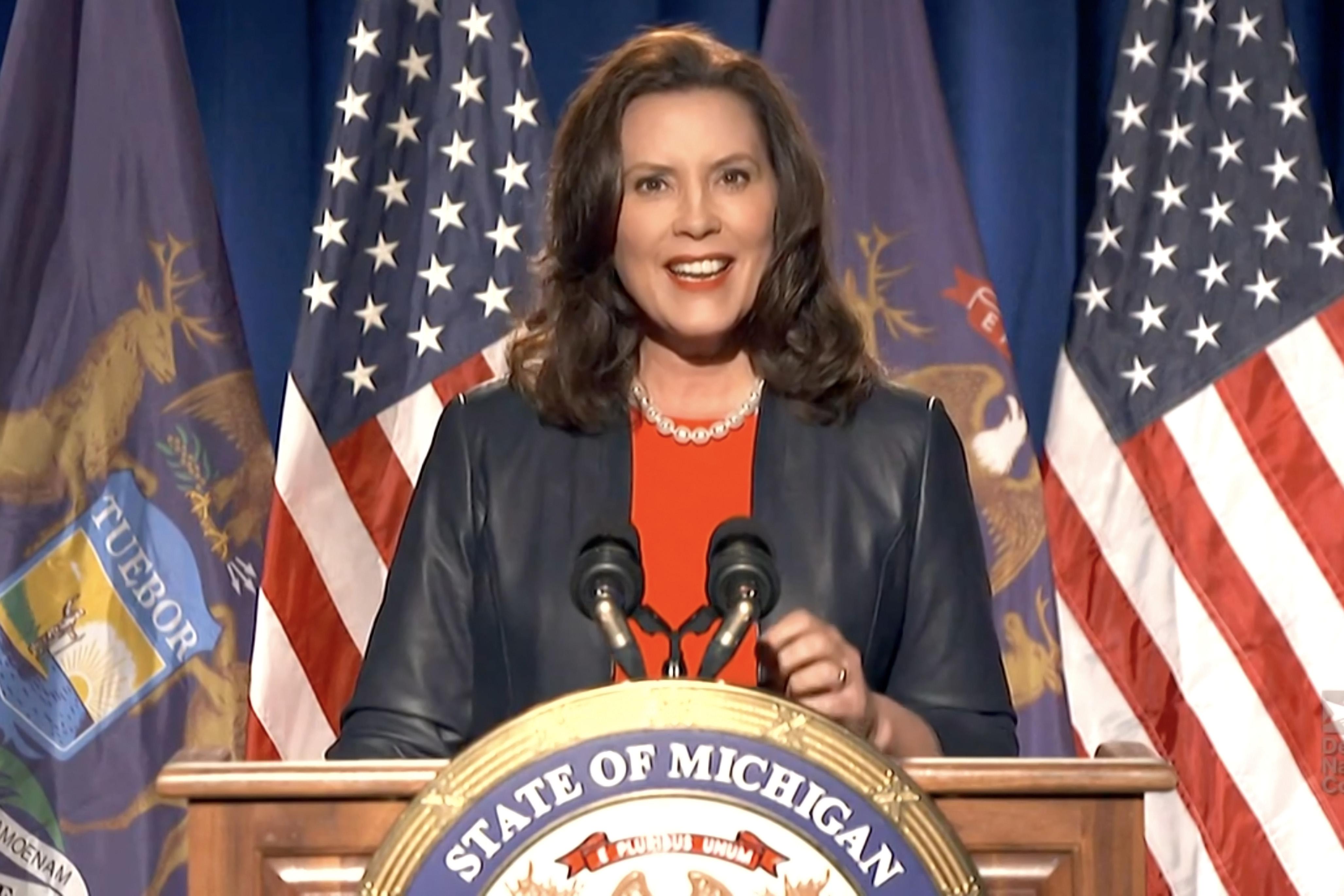 Whitmer, seen in a leather jacket, speaks at a podium bearing a Michigan seal.