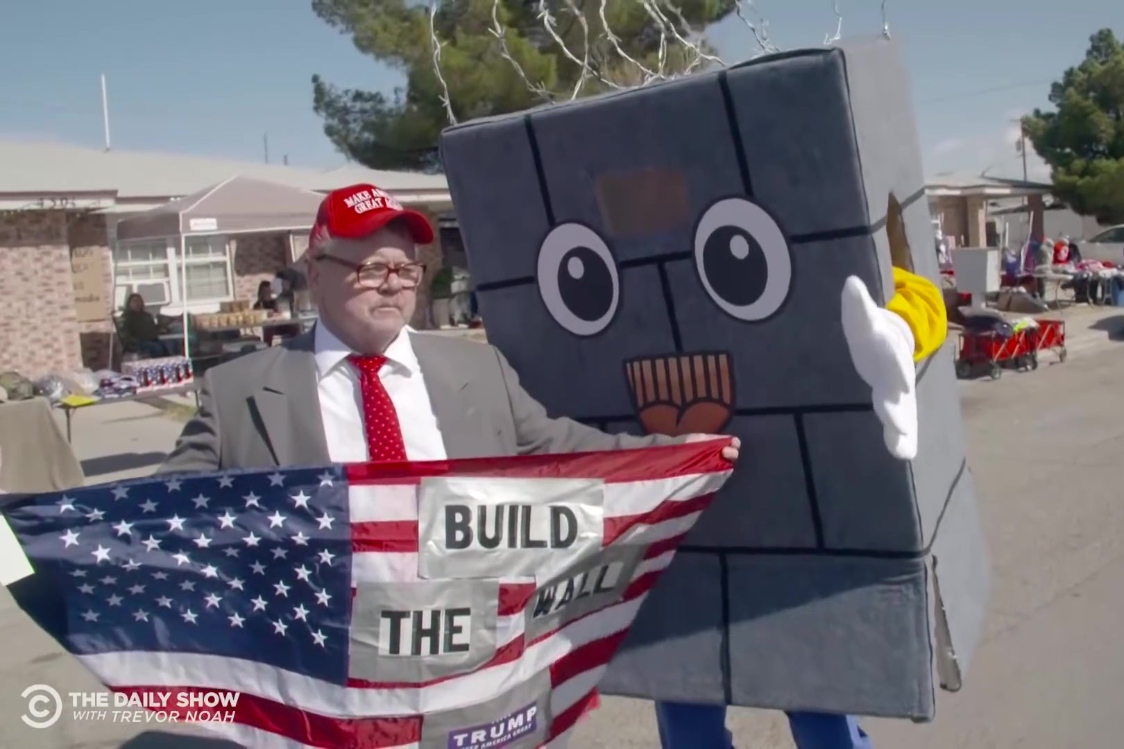 A man holding an American flag reading "Build the Wall" talks to a Daily Show correspondent dressed up as a cartoon wall.