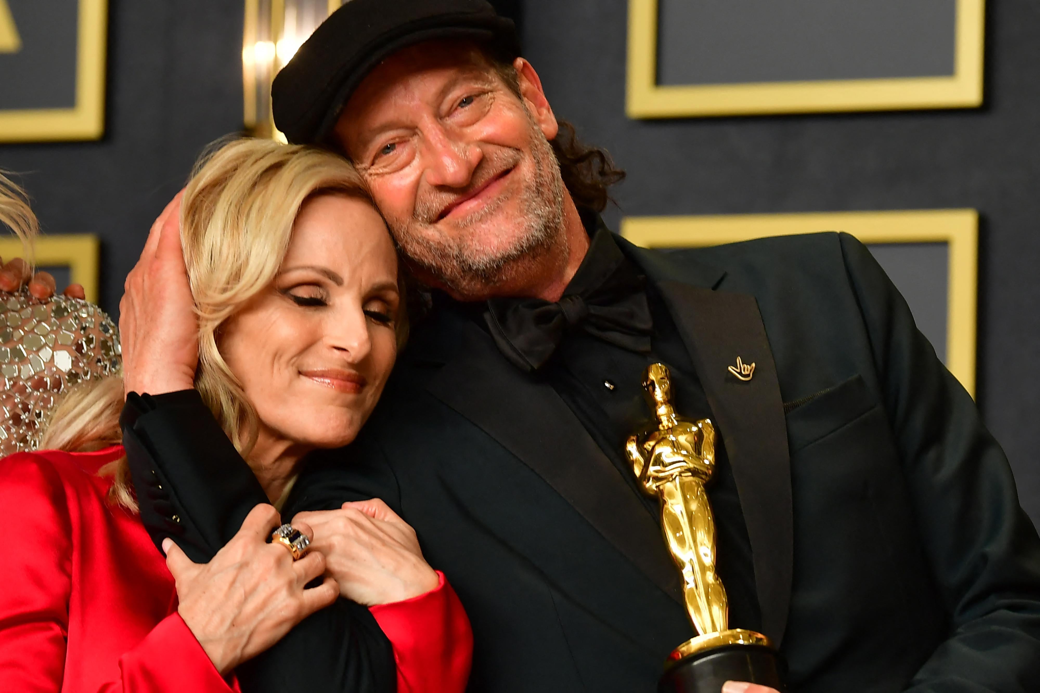 Troy Kotsur smiles at the camera as he holds up his Oscar statuette and embraces Marlee Matlin, who smiles and leans on his shoulder.