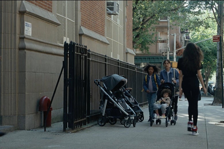 Rashida Jones pushes a stroller with a kid in it and a kid walking beside her down a city street