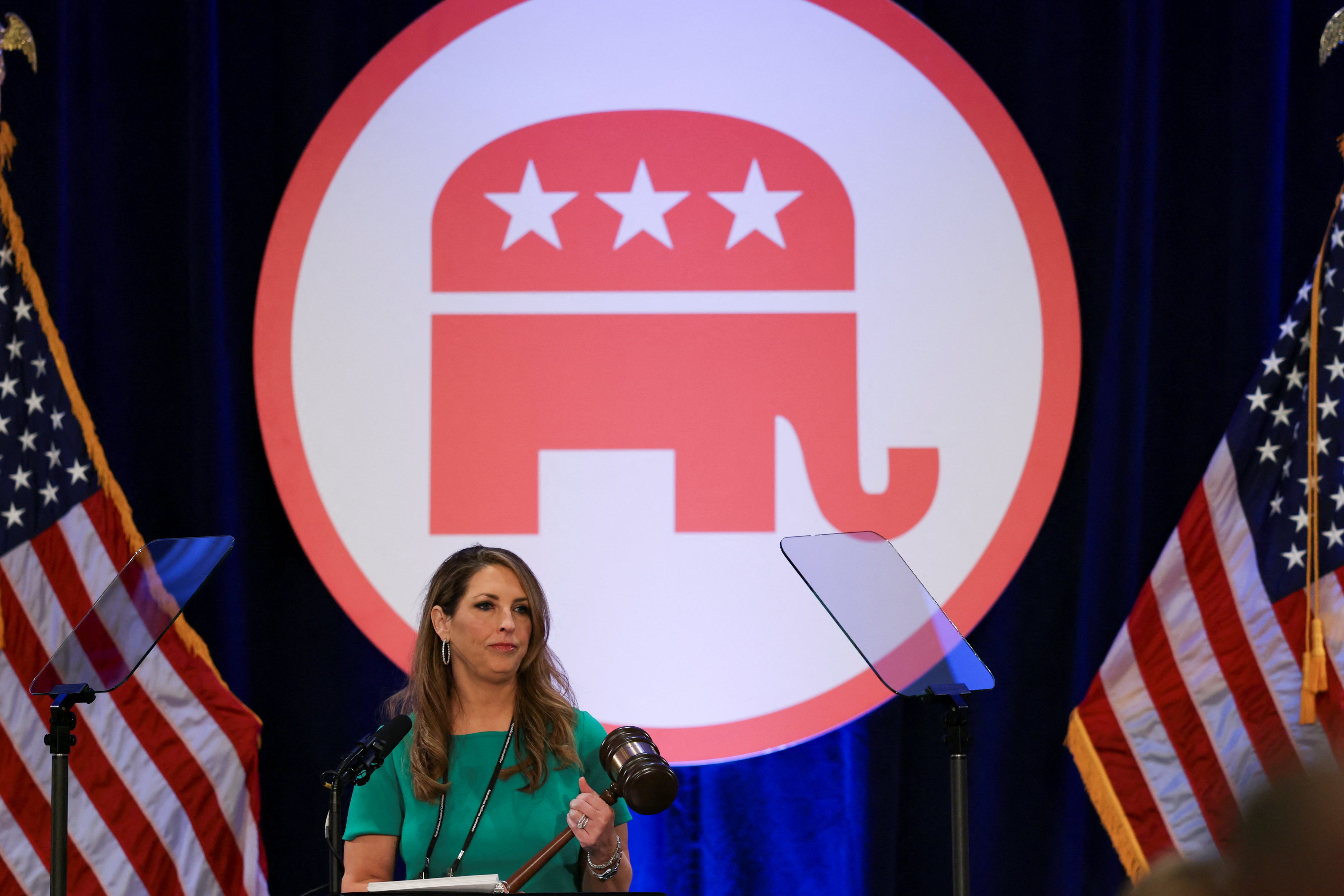 A woman holds a gavel with a large image of the GOP elephant behind her, flanked on either side by American flags.