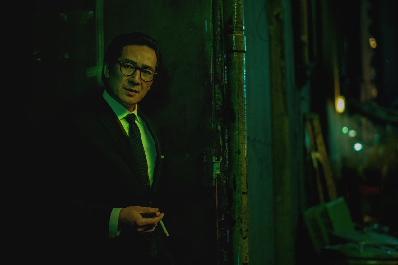 A man in a suit holds a cigarette in a neon green-lit alleyway.