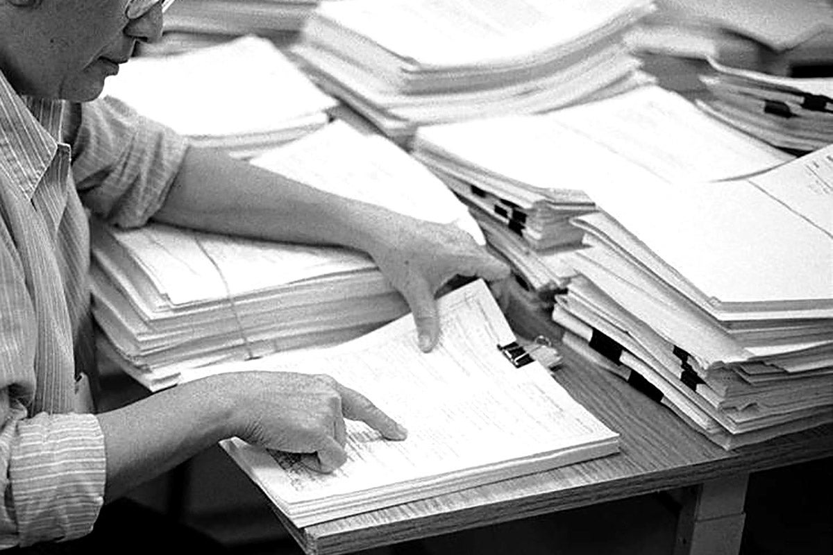 A person wearing glasses points at a particular line in a document on a desk piled heavily with thick documents.