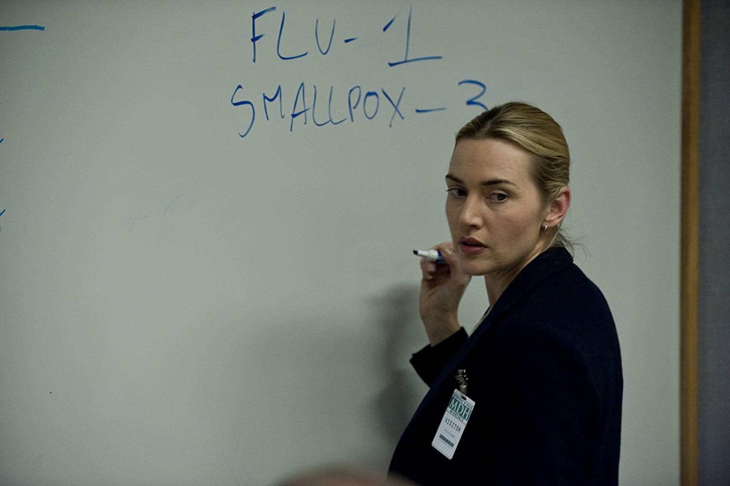 Kate Winslet holds a marker up in front of a whiteboard. The whiteboard says "FLU - 1, SMALLPOX - 3."